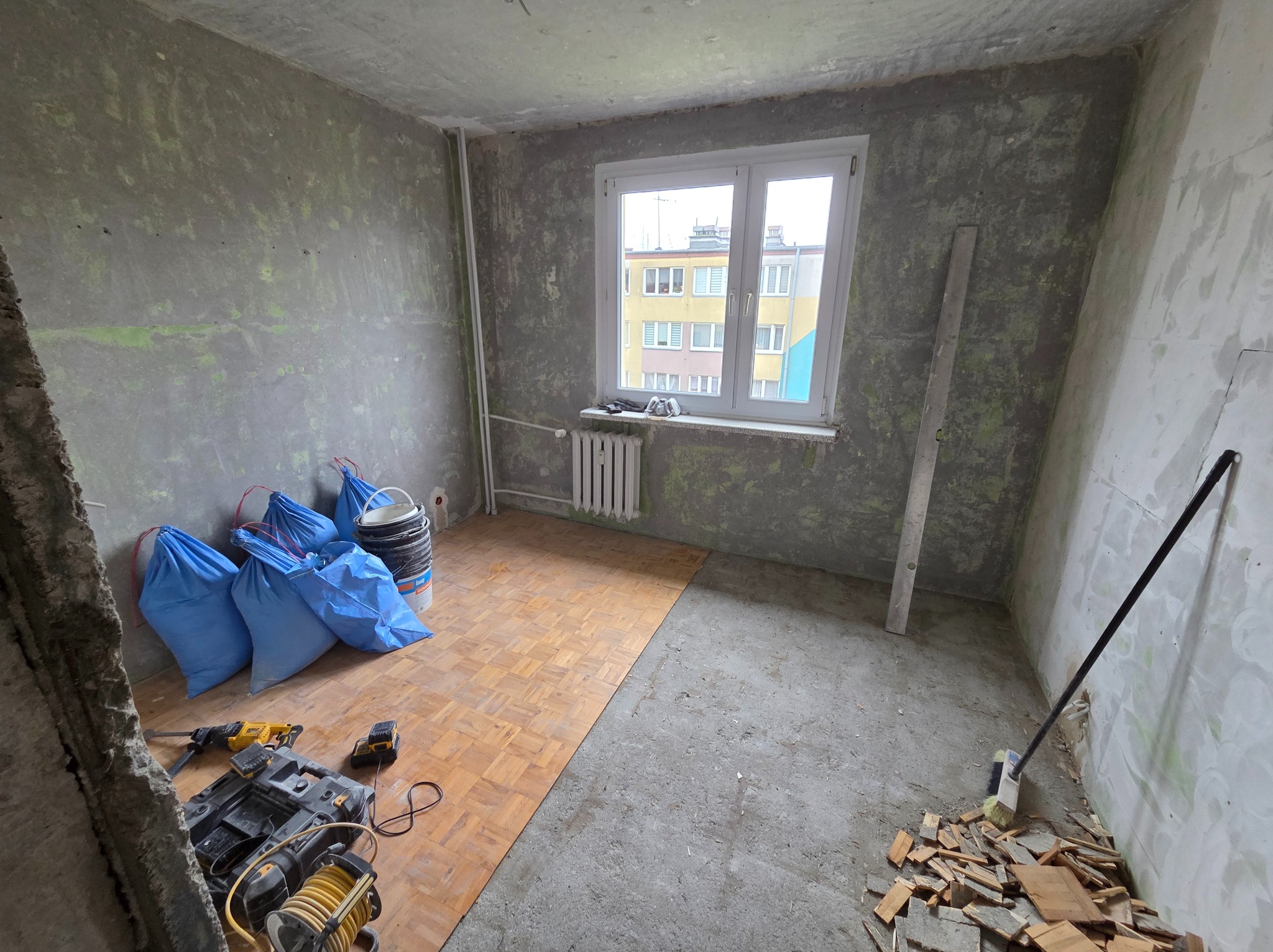 Pokój w trakcie remontu: częściowo ułożona drewniana podłoga, surowe ściany z resztkami farby, narzędzia i worki z odpadami budowlanymi. Widok na okno.