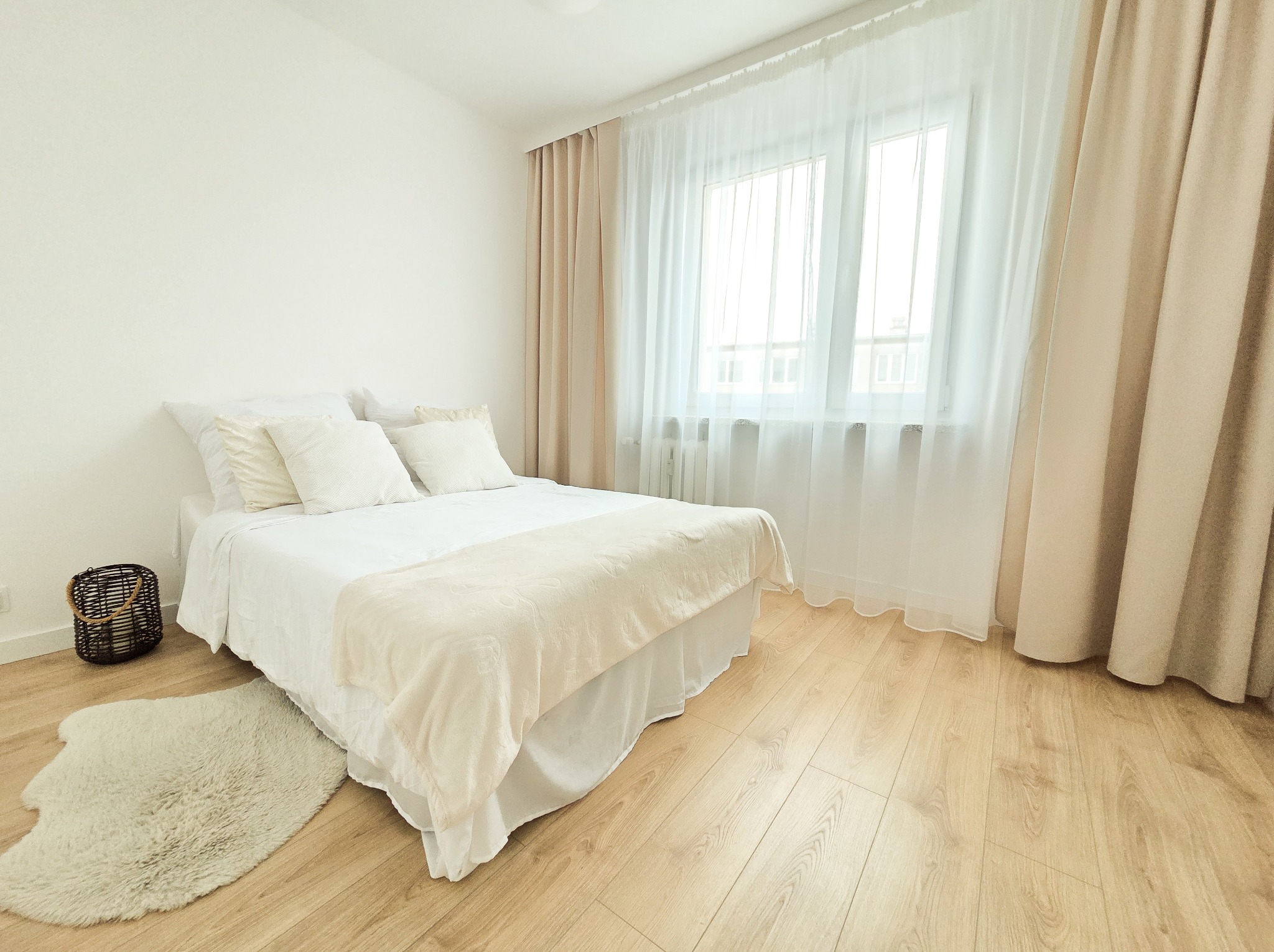 Jasna sypialnia z drewnianą podłogą, białym łóżkiem i oknem z beżowymi zasłonami. W rogu koszyk i dywanik. Minimalistyczny wystrój, ciepłe oświetlenie.