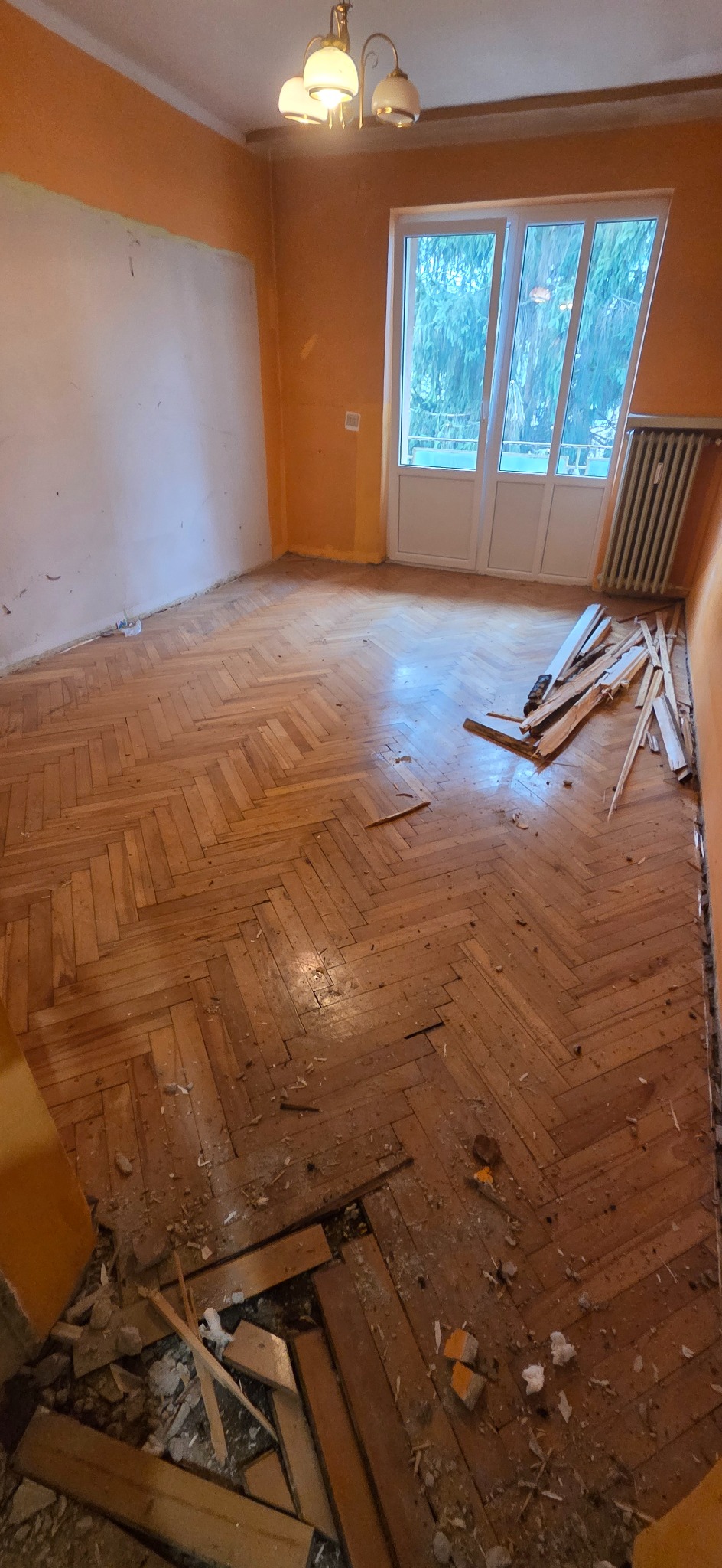 Pokój w trakcie demontażu parkietu ułożonego w jodełkę. Widoczne fragmenty starej podłogi, odspojone deski i resztki kleju. Pomarańczowe ściany i okno balkonowe w tle.