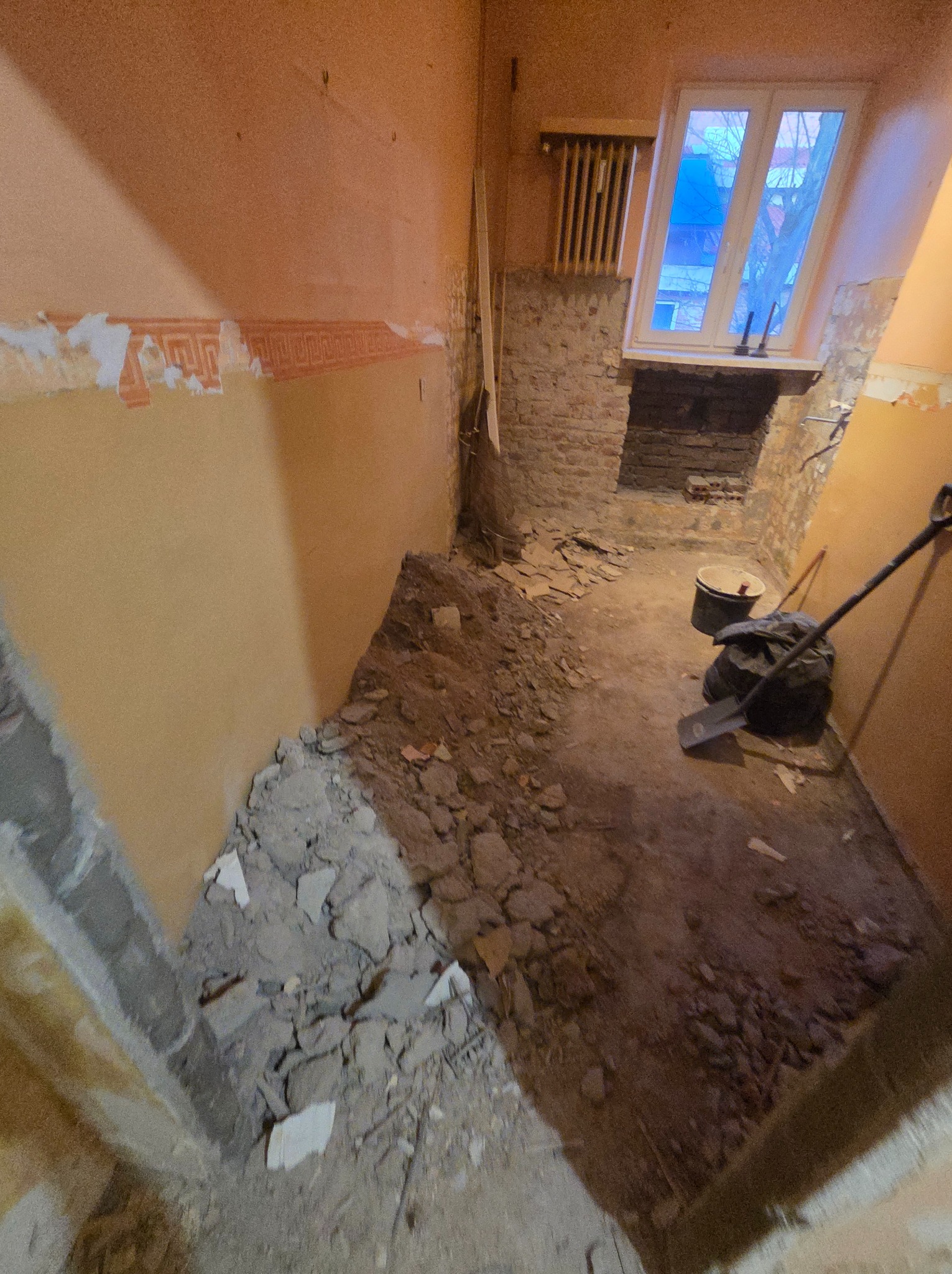 Wnętrze w trakcie demontażu: fragmenty ścian, gruz na podłodze, widoczne instalacje. Przy oknie wiadro i łopata. Przygotowanie do remontu w pomieszczeniu.