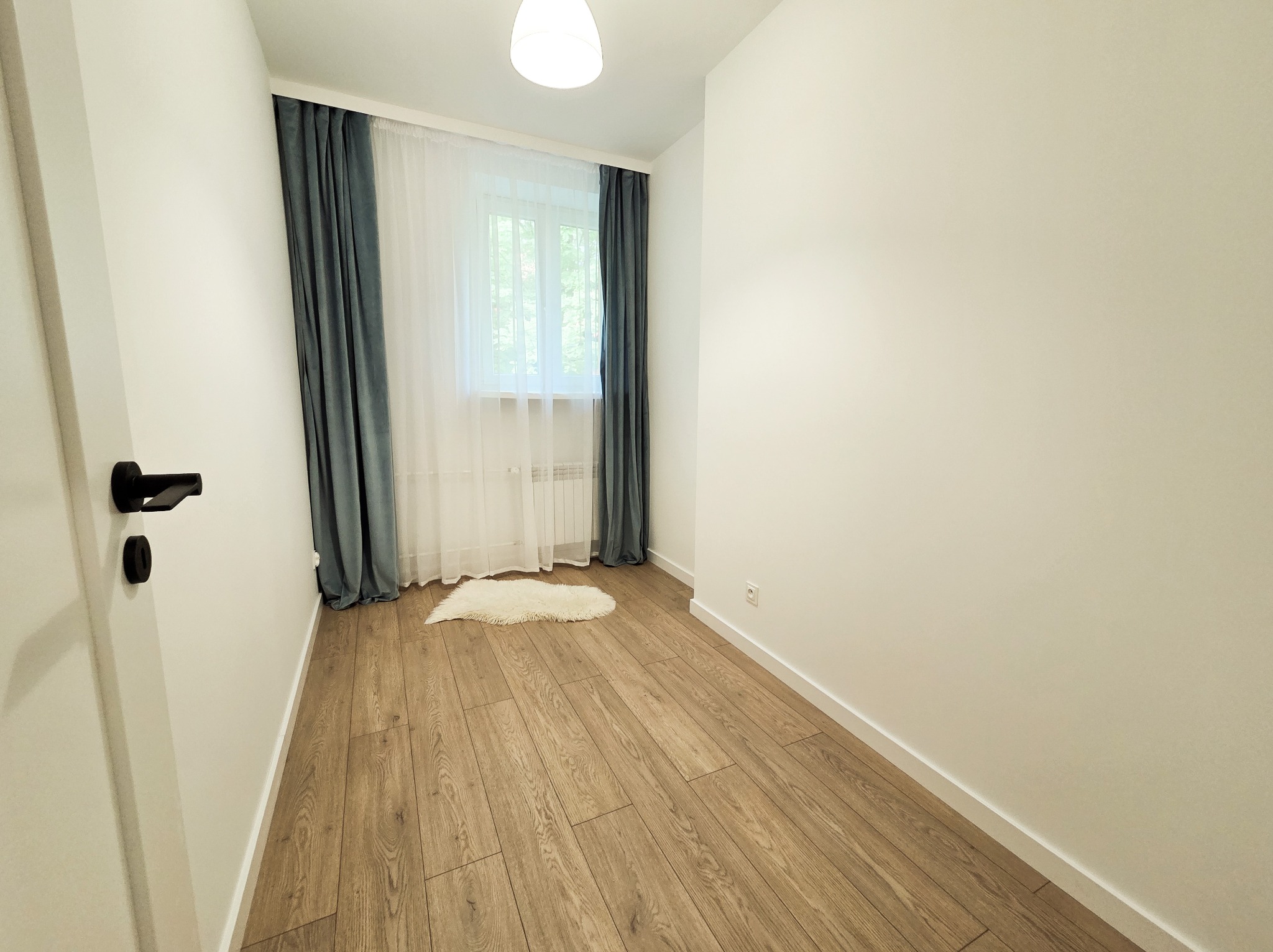Jasne wnętrze pokoju po remoncie: drewniana podłoga, białe ściany, okno z zasłonami i mały dywanik. Widoczna klamka drzwi.