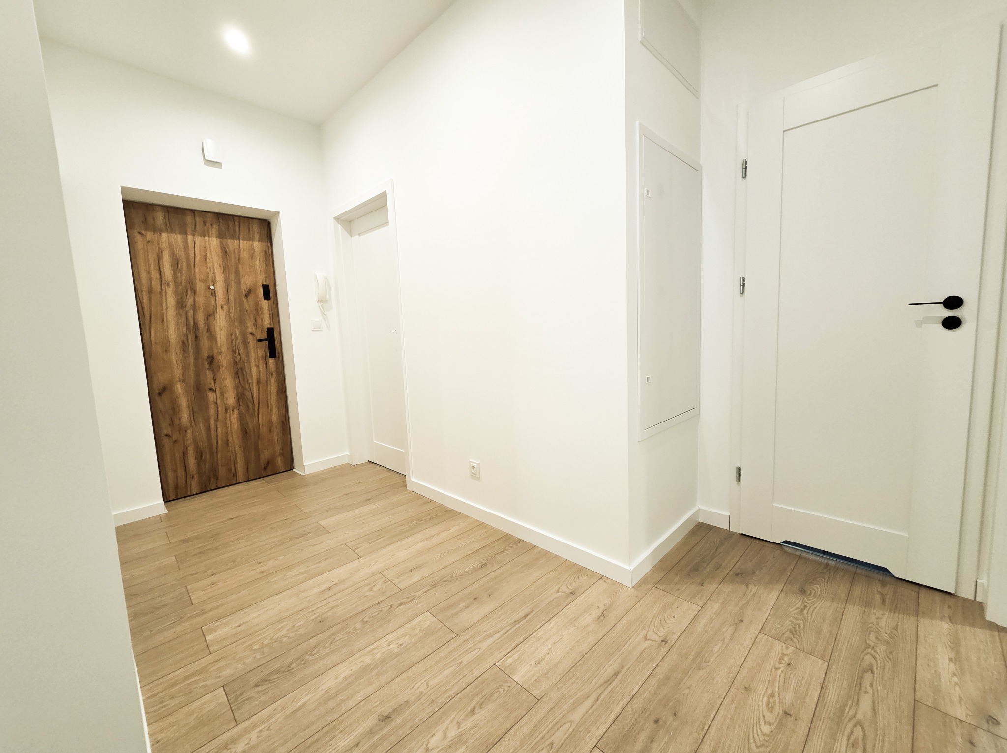 Jasne panele podłogowe w korytarzu z dwojgiem drzwi: drewnianymi wejściowymi i białymi wewnętrznymi. Minimalistyczny design, białe ściany i listwy przypodłogowe.