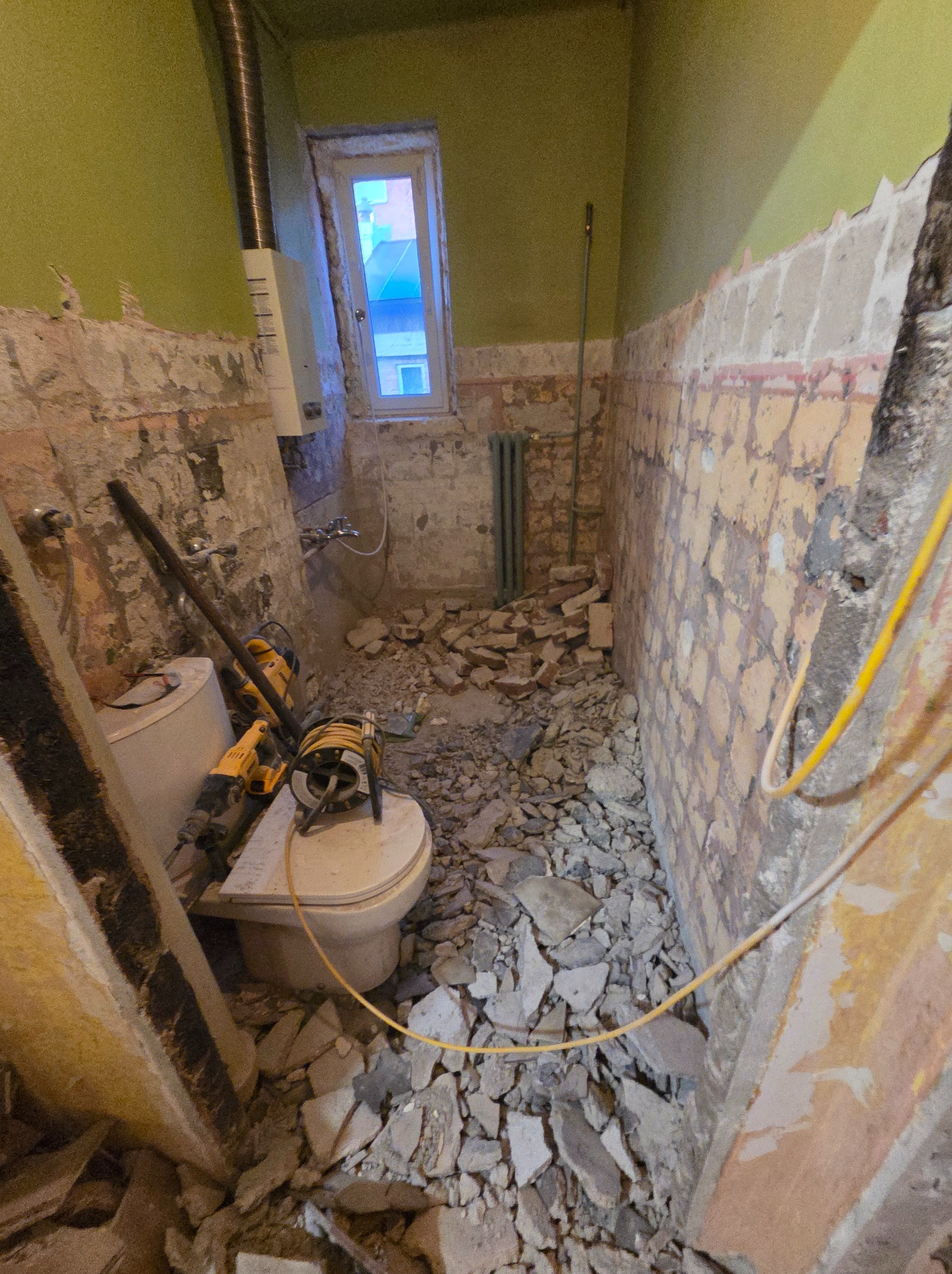 Demontaż starej łazienki: gruz, narzędzia (wiertarka, młot udarowy) i fragmenty ścian z odsłoniętą cegłą. Widoczne okno i bojler. Wnętrze w trakcie prac remontowych.