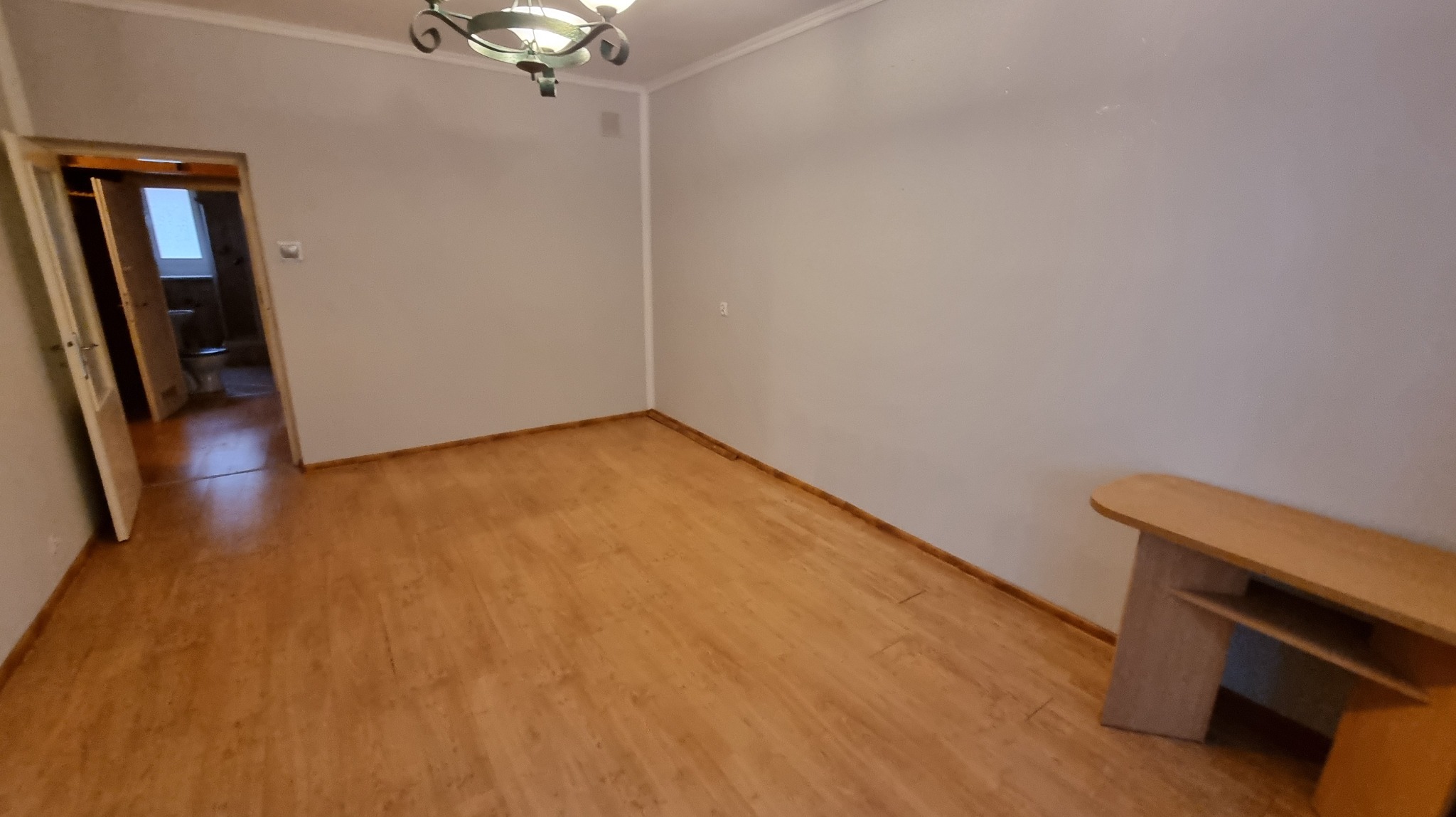Pusty pokój z drewnianą podłogą i szarymi ścianami. W tle otwarte drzwi do łazienki. Widoczny fragment mebla i ozdobna lampa sufitowa.