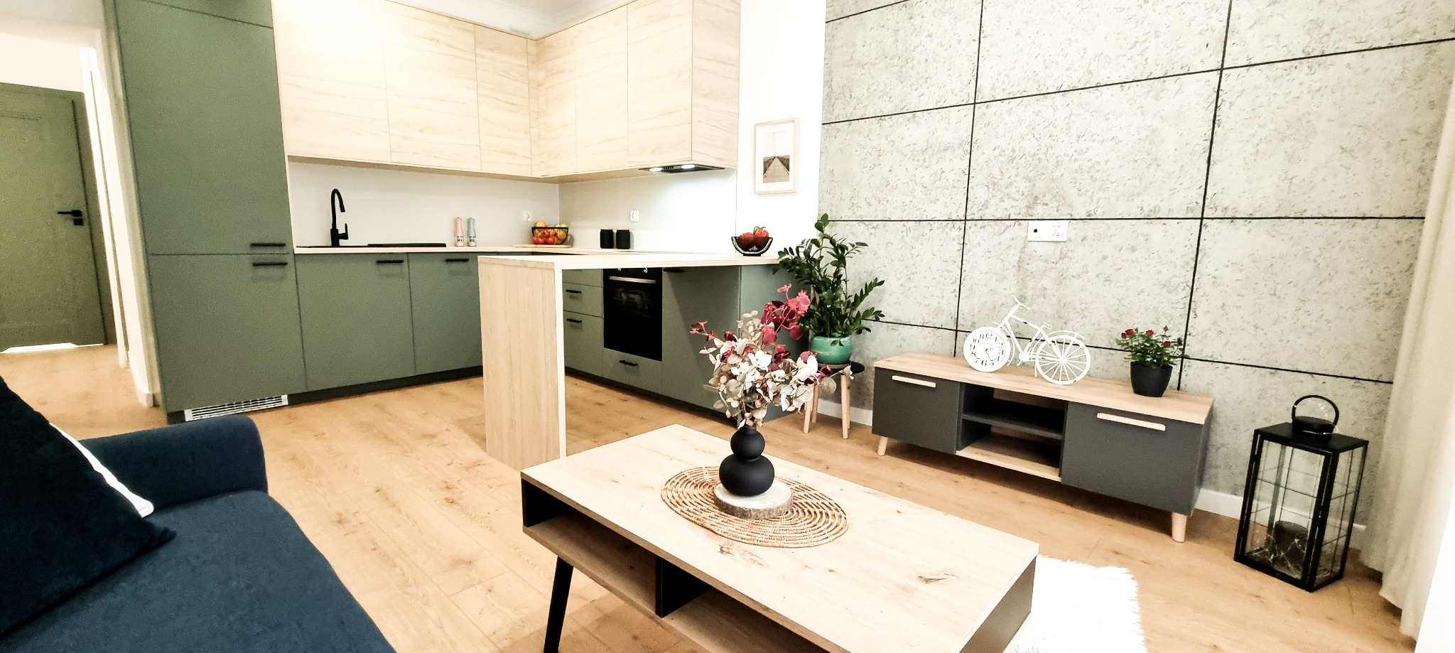 Nowoczesne wnętrze: kuchnia z zielonymi szafkami i drewnianym blatem, połączona z salonem. Jasna podłoga, szara ściana z dekoracyjnymi panelami. Minimalistyczny design.