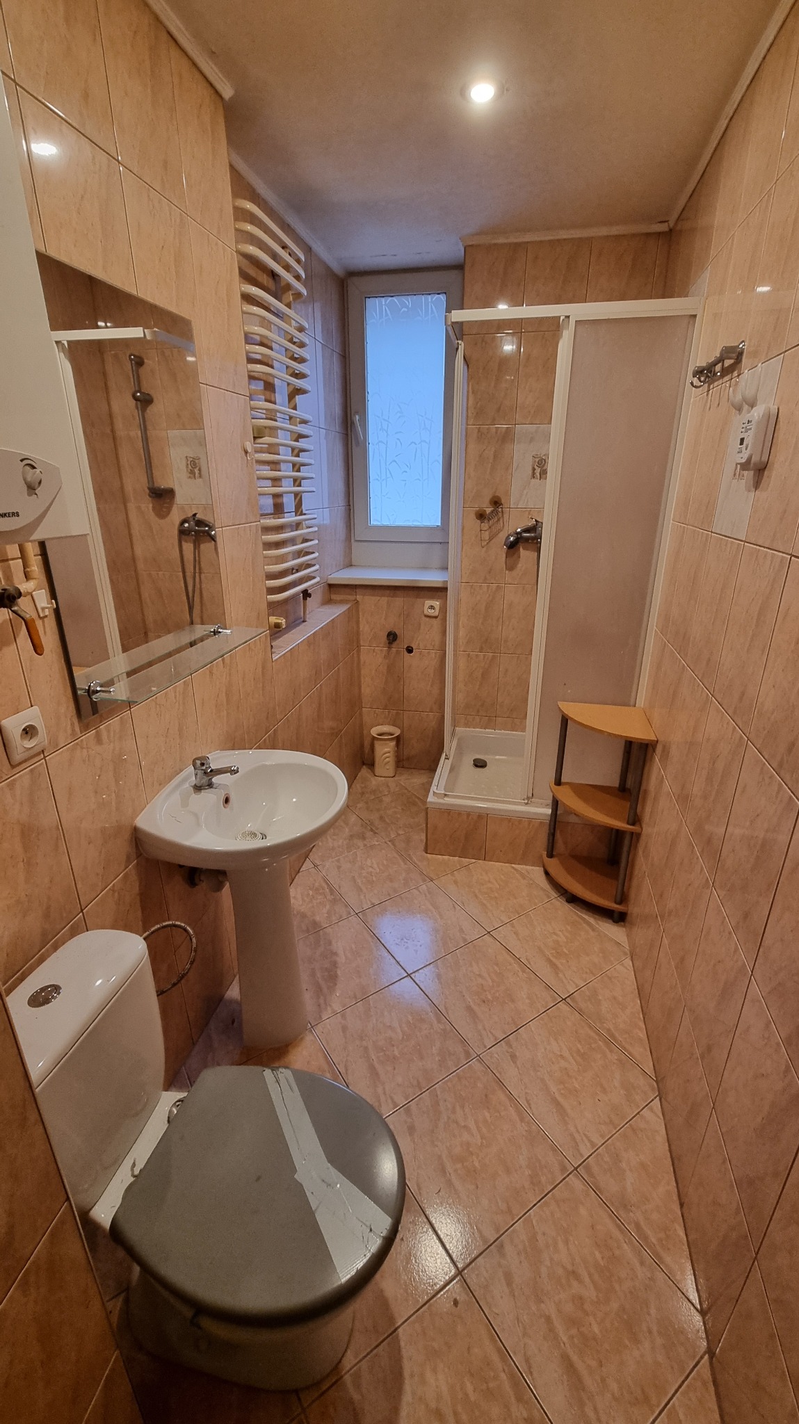 Łazienka w beżowych płytkach z kabiną prysznicową, umywalką na postumencie i toaletą z uszkodzoną deską. Grzejnik drabinkowy przy oknie. Widok z perspektywy.