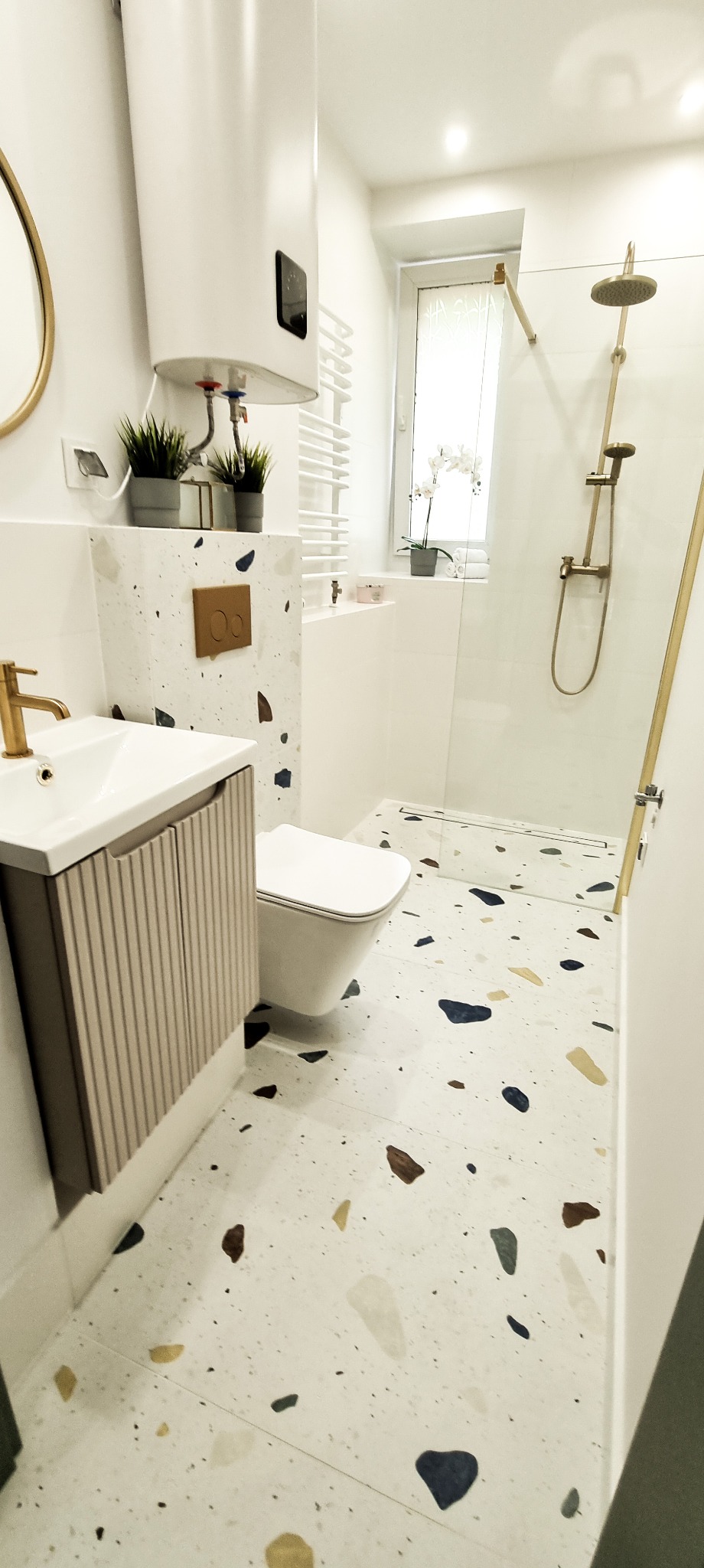 Nowoczesna łazienka z terazzo: białe ściany, wisząca toaleta, szafka z umywalką i złota armatura. Minimalistyczny design z roślinami doniczkowymi.