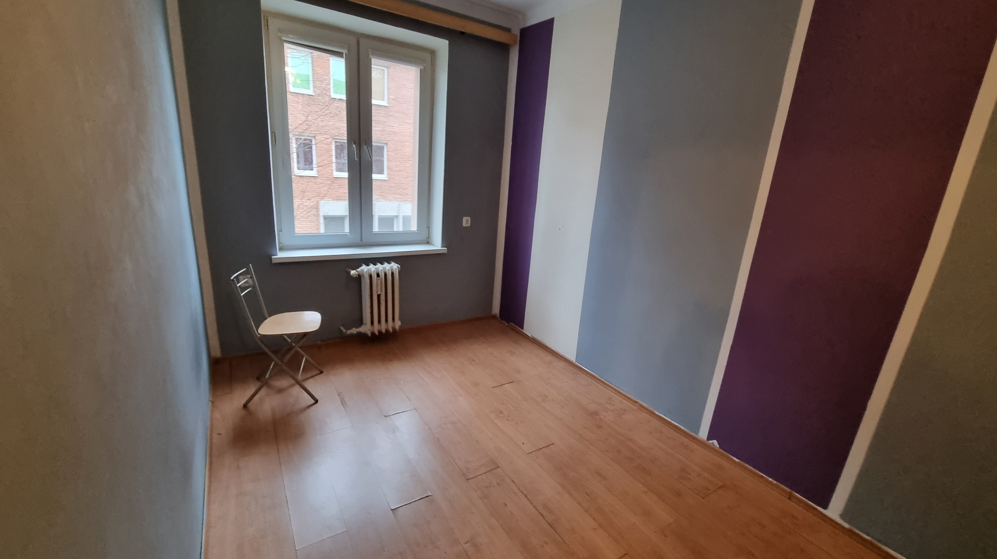 Pomalowane ściany w pokoju w pasy: fioletowy, szary i biały. Na podłodze panele. Przy oknie krzesło i grzejnik. Widok na budynek z zewnątrz.