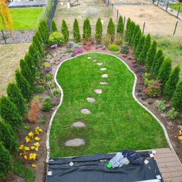 ogrody.filip - Widok z góry na świeżo założony trawnik z ścieżką z kamieni, obrzeżami i nasadzeniami z tuj. W tle domy jednorodzinne i przygotowane podłoże pod dalsze prace.