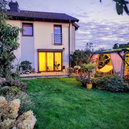 ogrody.filip - Ogród z domem w tle o zmierzchu: wypielęgnowany trawnik, kwitnące hortensje, hamak w altanie i oświetlony dom z tarasem. Romantyczny nastrój.