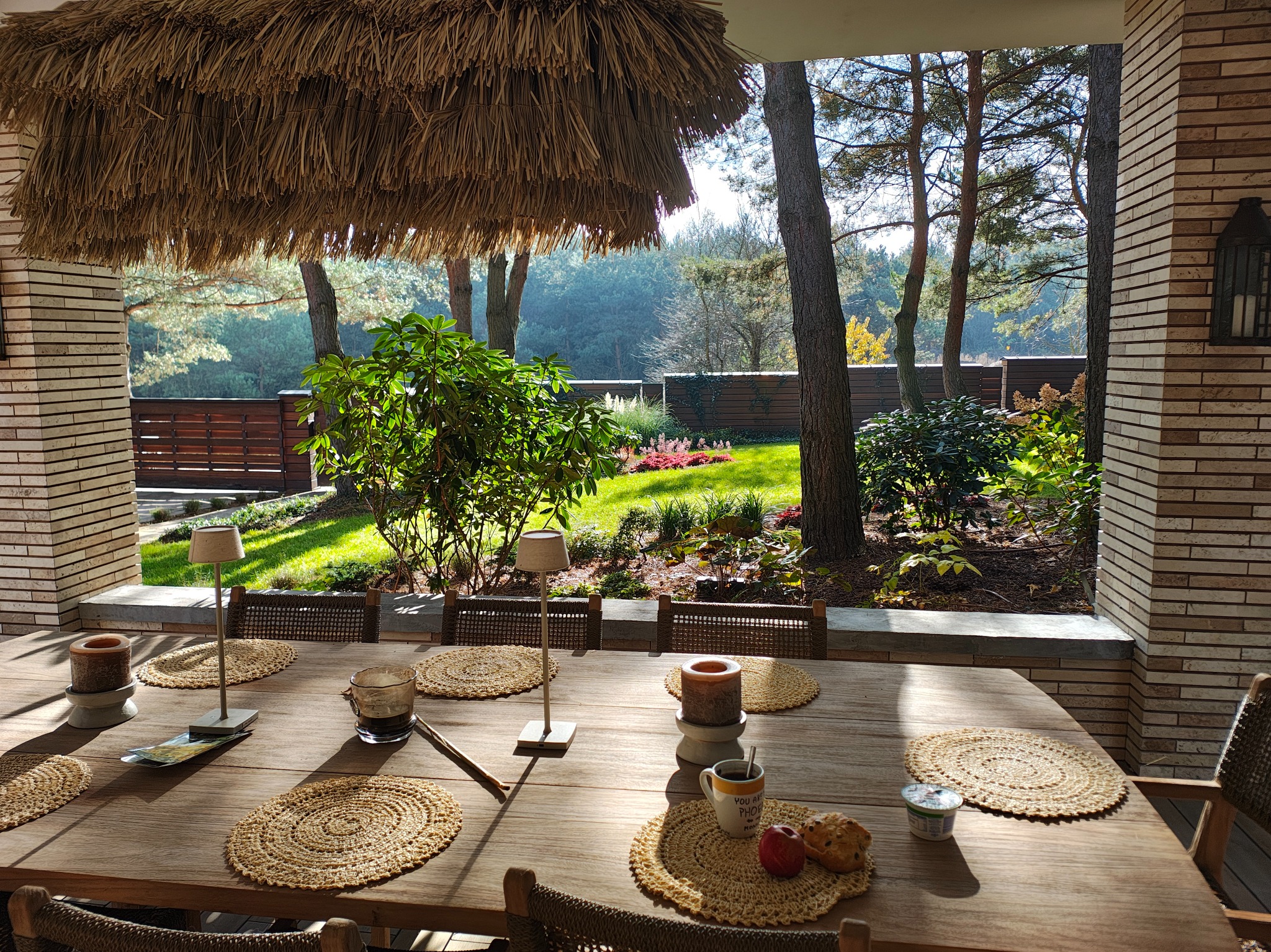 Zadaszony taras z drewnianym stołem i widokiem na zadbany ogród z drzewami i krzewami. Dekoracyjne podkładki na stole i lampki tworzą przytulną atmosferę.