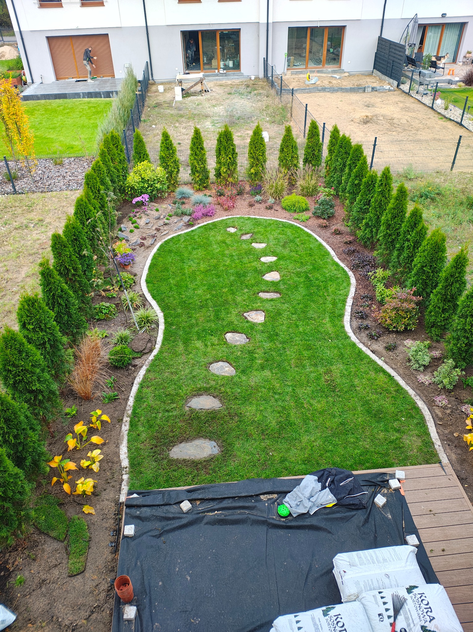 Widok z góry na świeżo założony trawnik z ścieżką z kamieni, obrzeżami i nasadzeniami z tuj. W tle domy jednorodzinne i przygotowane podłoże pod dalsze prace.