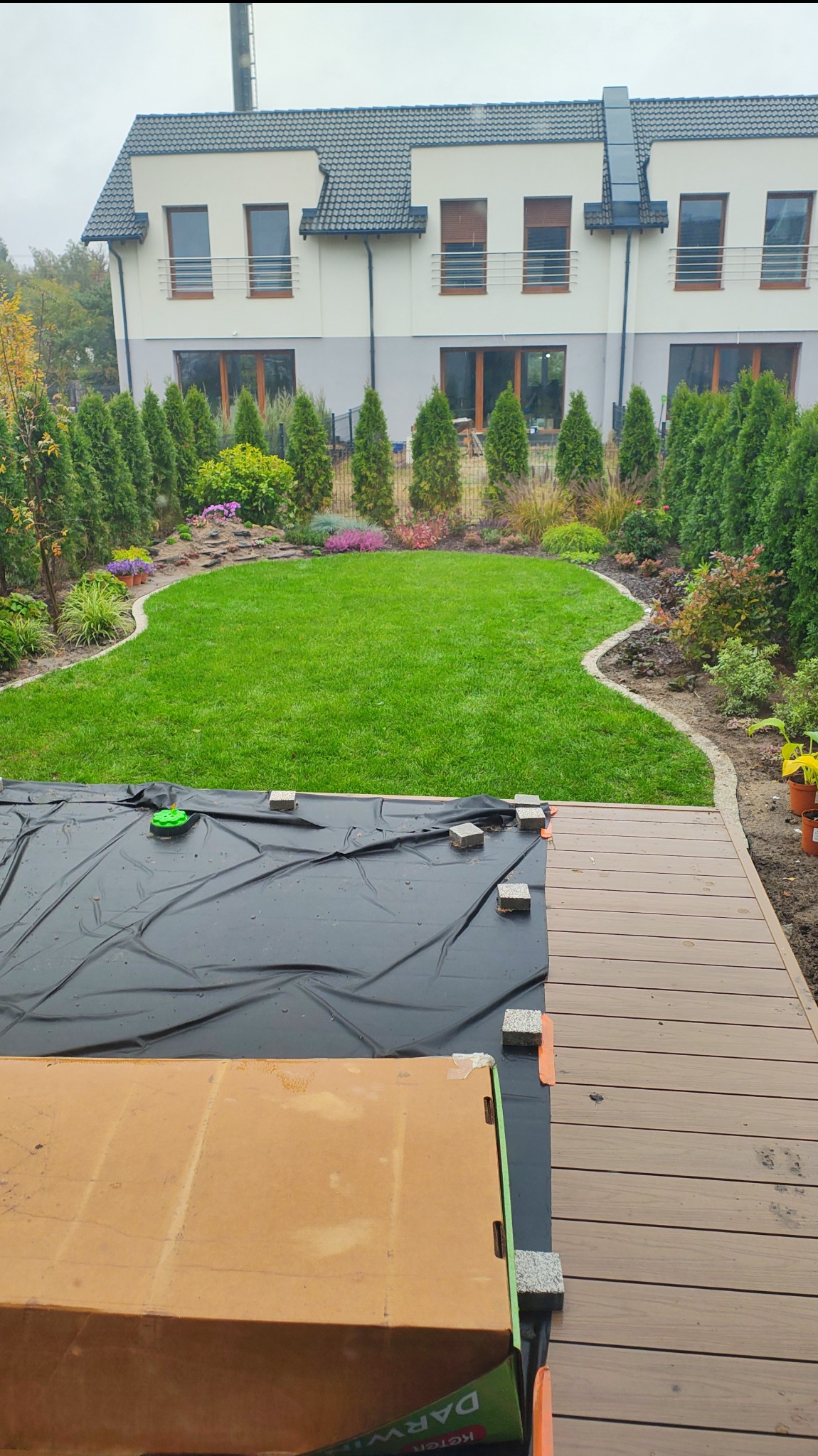 Widok na trawnik z rabatami kwiatowymi i tujami, z fragmentem budowy tarasu z desek kompozytowych i folii ochronnej na pierwszym planie. Ujęcie z góry.