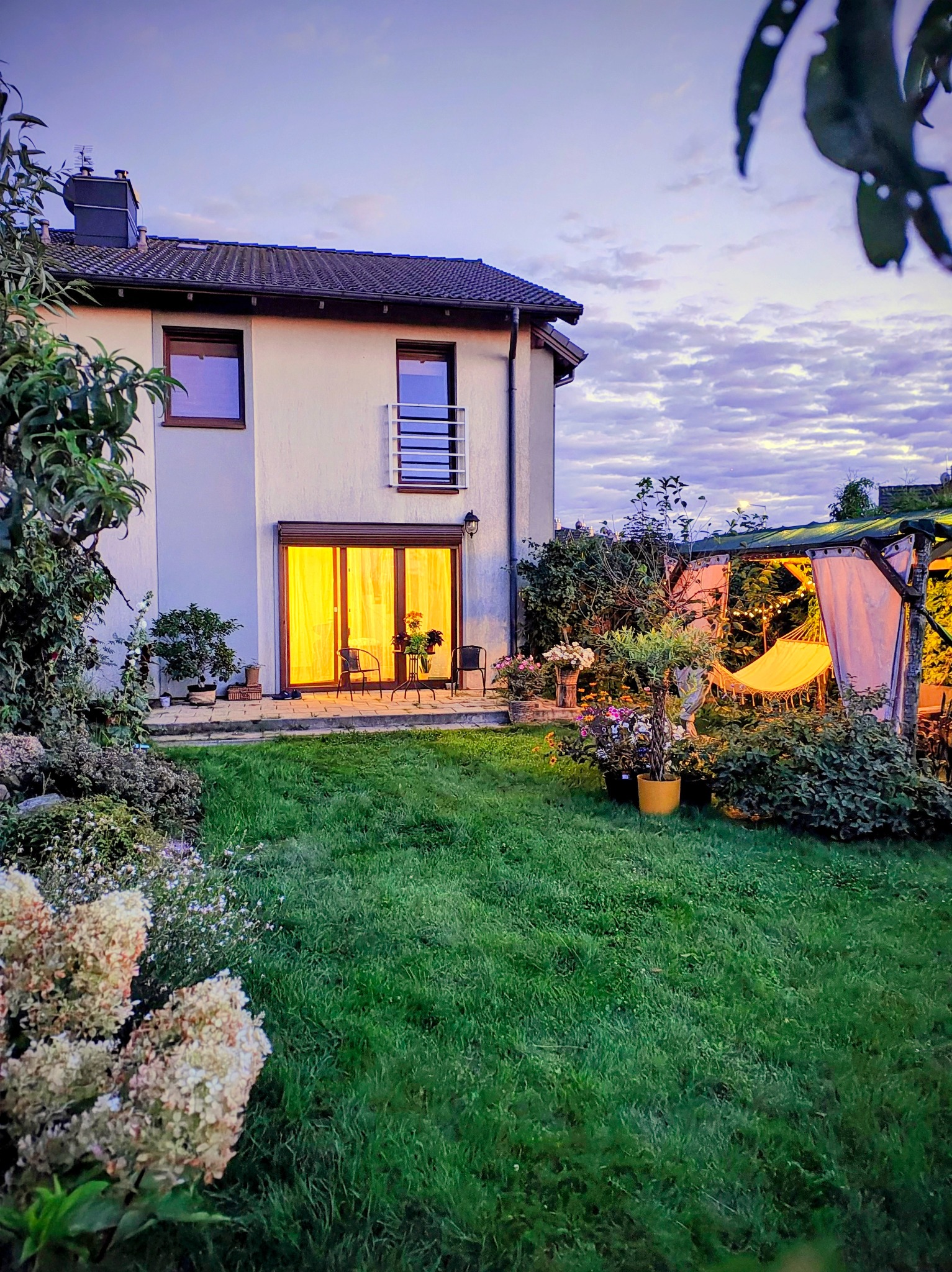 Ogród z domem w tle o zmierzchu: wypielęgnowany trawnik, kwitnące hortensje, hamak w altanie i oświetlony dom z tarasem. Romantyczny nastrój.