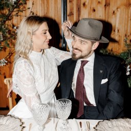 MORGAN & NESS - Elegancka para przy stole: kobieta w koronkowej sukience dotyka kapelusza mężczyzny w ciemnym garniturze i bordowym krawacie. Romantyczna atmosfera, drewniane tło.