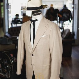 MORGAN & NESS - Beżowy garnitur na manekinie z kapeluszem i krawatem bolo, w tle stylowe wnętrze z motocyklem. Detal krawiecki, elegancja, szycie na miarę.