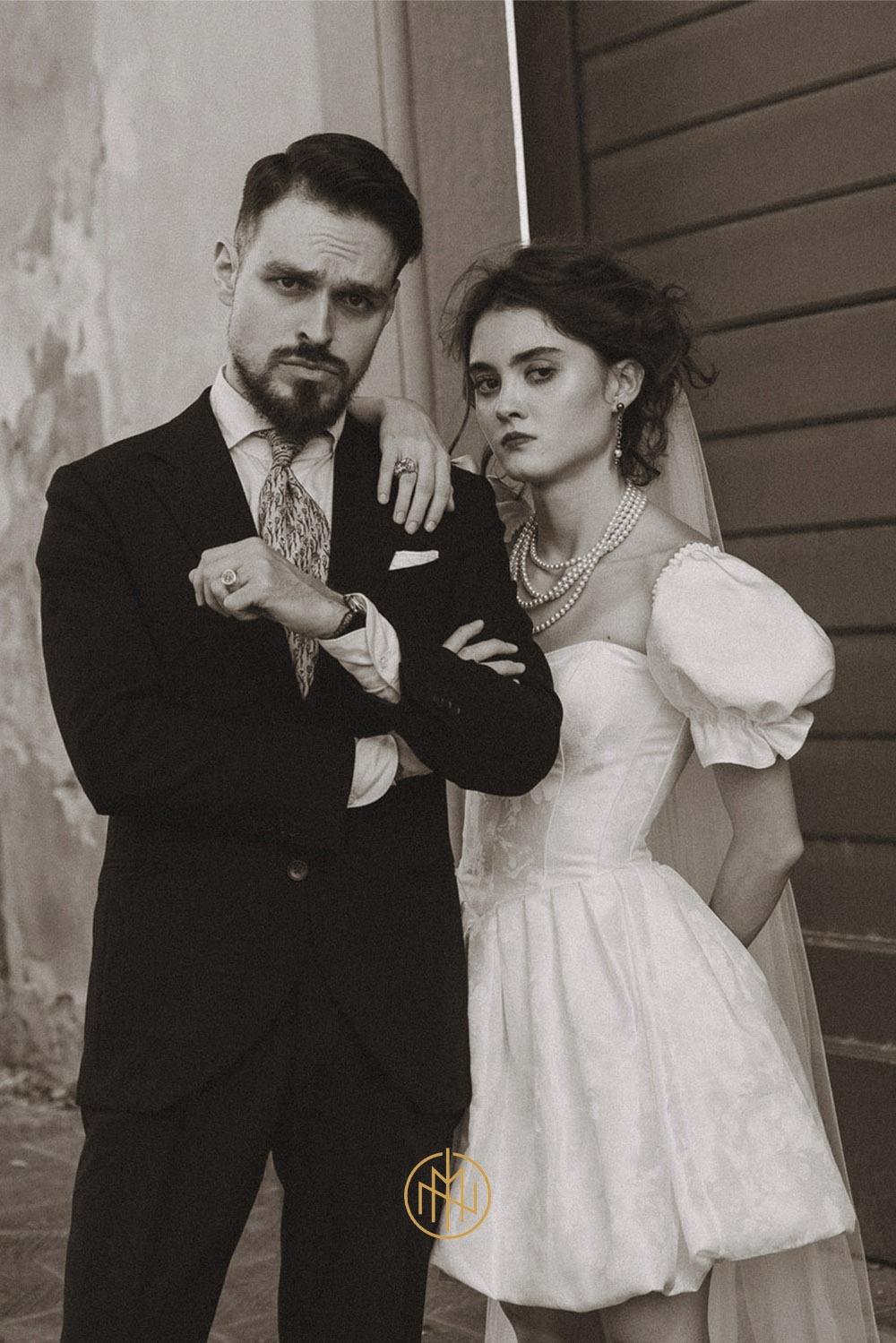 Sepiowy portret pary: mężczyzna w ciemnym garniturze z wzorzystym krawatem, kobieta w białej sukni ślubnej z perłami, oboje z poważnym wyrazem twarzy na tle ściany.