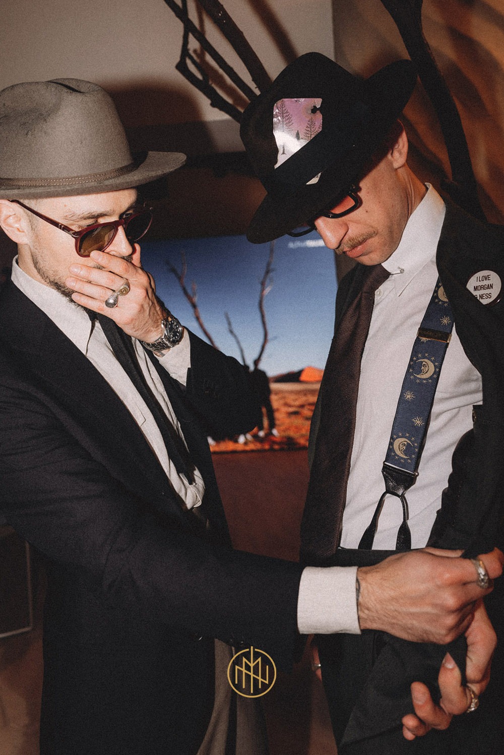 Dwóch mężczyzn w eleganckich strojach: jeden w szarym kapeluszu i garniturze, drugi w czarnym kapeluszu, koszuli i szelkach z motywem księżyca. Nietypowe stylizacje.