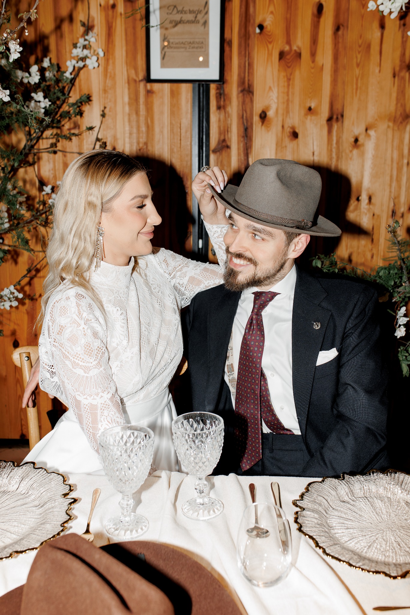 Elegancka para przy stole: kobieta w koronkowej sukience dotyka kapelusza mężczyzny w ciemnym garniturze i bordowym krawacie. Romantyczna atmosfera, drewniane tło.