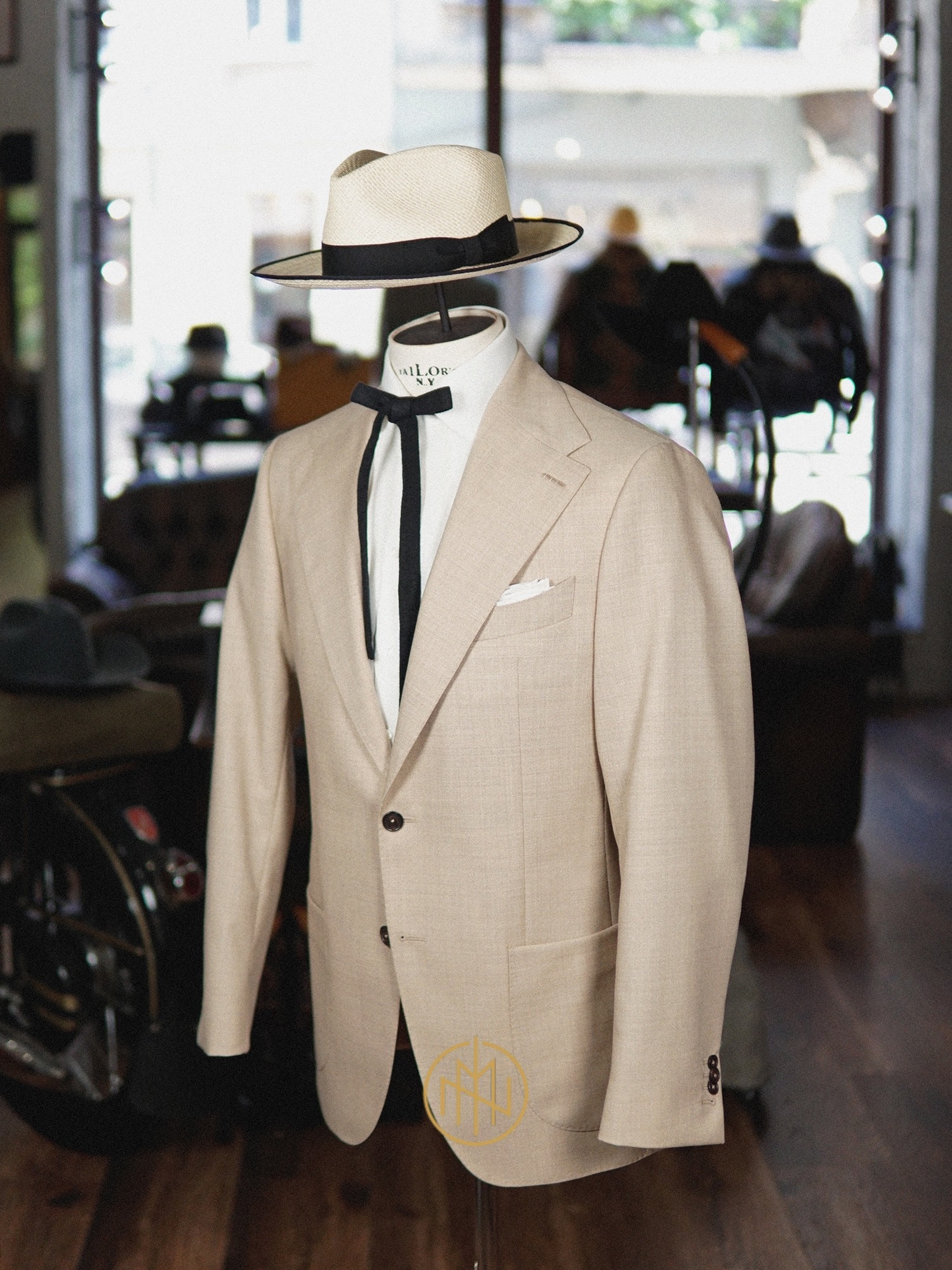 Beżowy garnitur na manekinie z kapeluszem i krawatem bolo, w tle stylowe wnętrze z motocyklem. Detal krawiecki, elegancja, szycie na miarę.