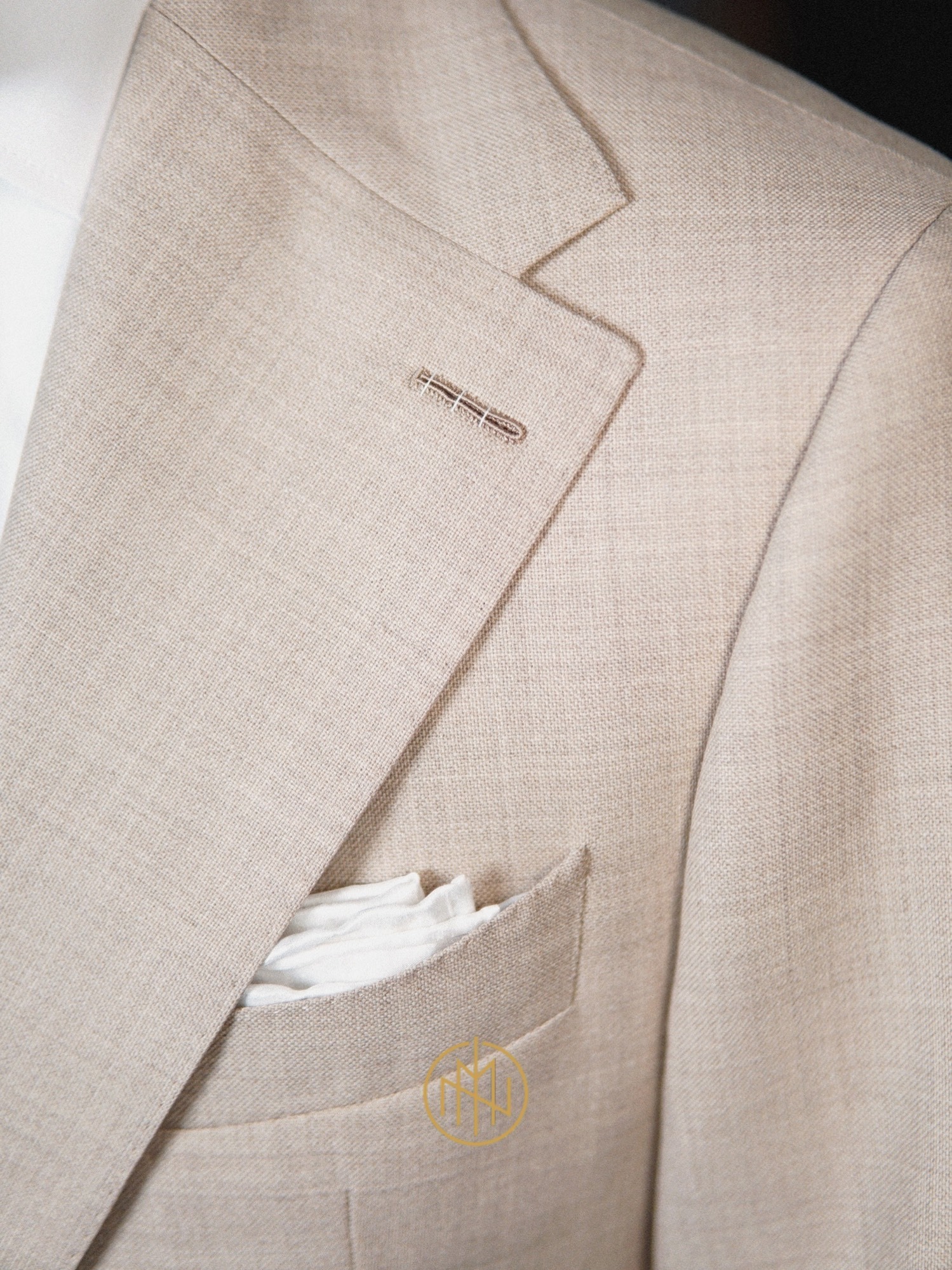 Beżowa marynarka z lnu, widoczny fragment klapy, kieszeń z białą poszetką i delikatny detal w butonierce. Elegancki krój i naturalna faktura materiału.