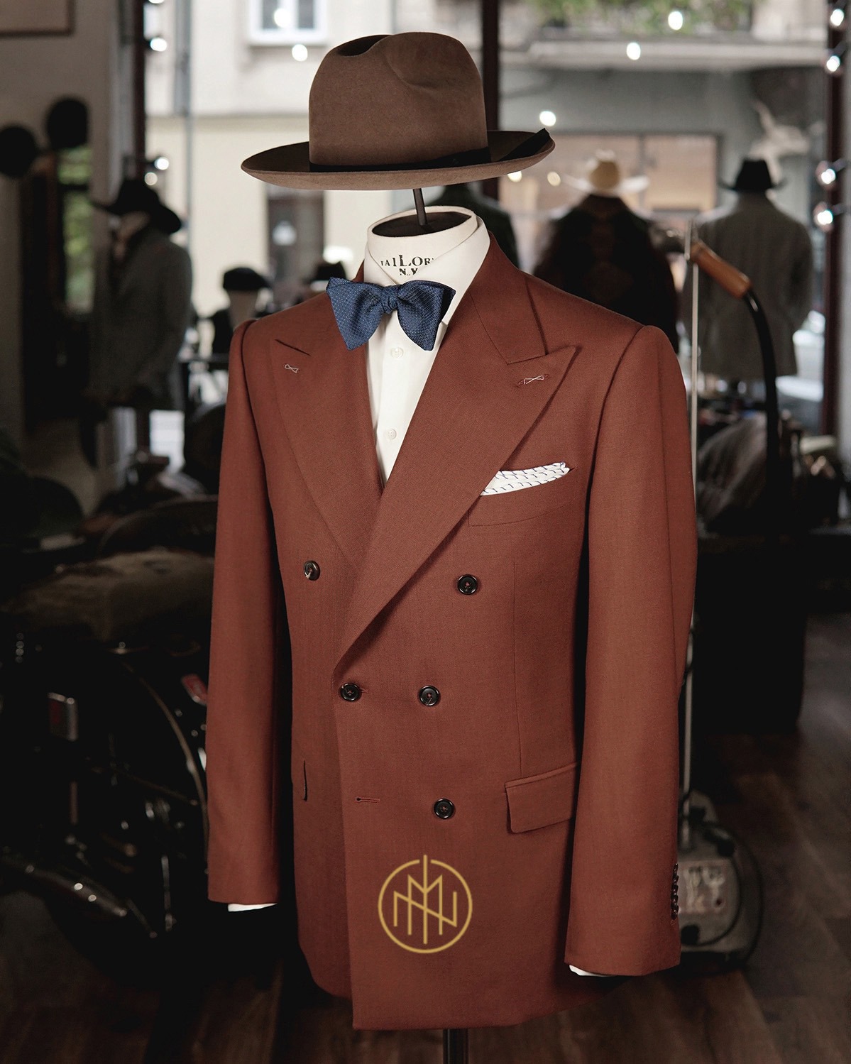 Brązowy, dwurzędowy blezer z muchą i kapeluszem na manekinie w pracowni krawieckiej. Widoczne detale: logo na klapie, wzorzysta poszetka, ciemne guziki.