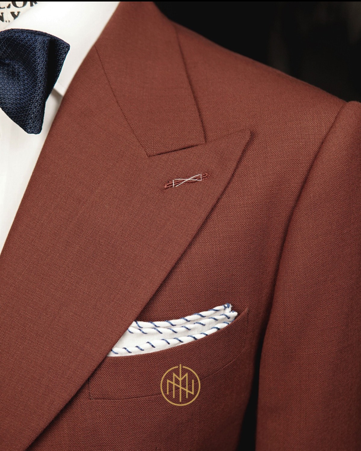 Brązowy, teksturowany blezer z poszetką w kieszonce, granatowa mucha, ozdobna szpilka w klapie i subtelne logo na kieszeni. Elegancki detal.