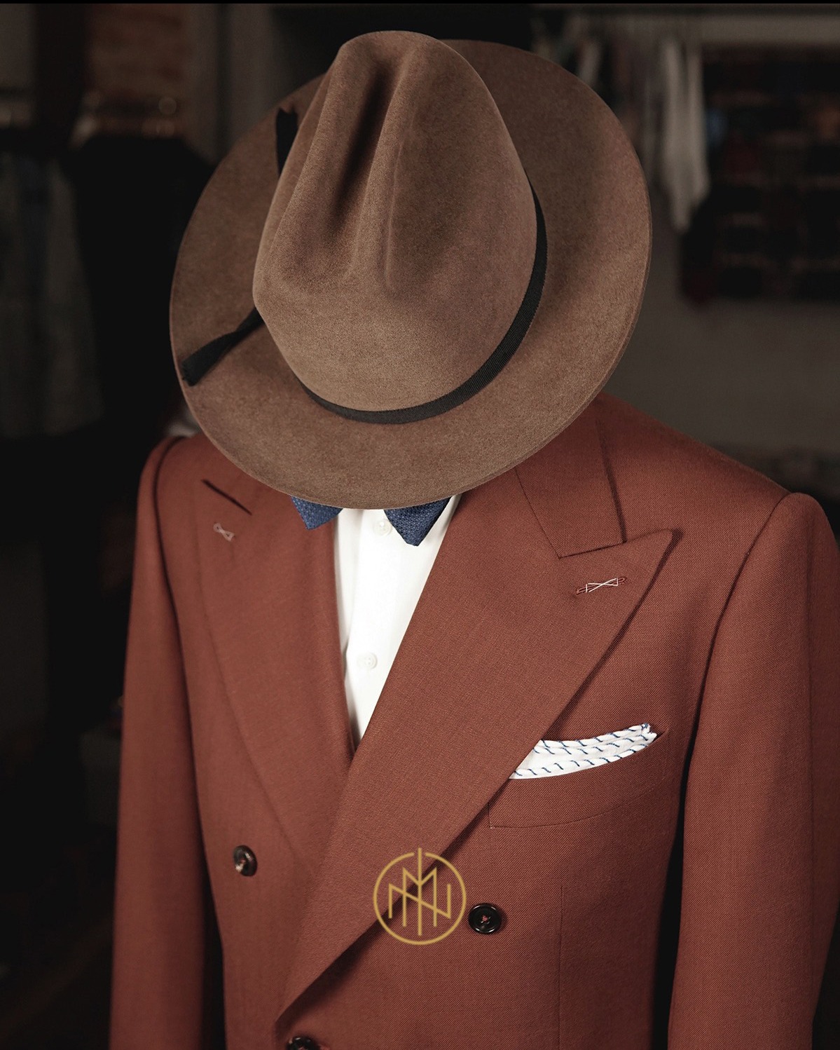 Brązowy, dwurzędowy garnitur z muchą i poszetką, nakryty brązowym kapeluszem. Elegancki strój na manekinie w krawieckim atelier.