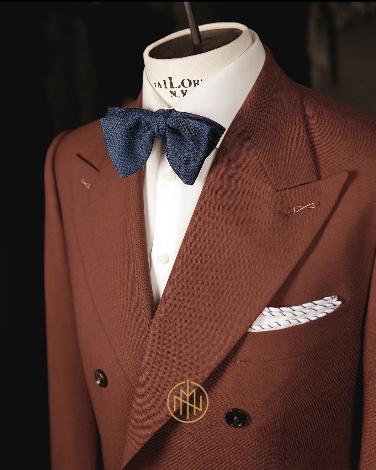 Brązowy, dwurzędowy garnitur z granatową muchą i poszetką, prezentowany na manekinie krawieckim z logo 'Tailor N.Y.', w eleganckim, stonowanym oświetleniu.
