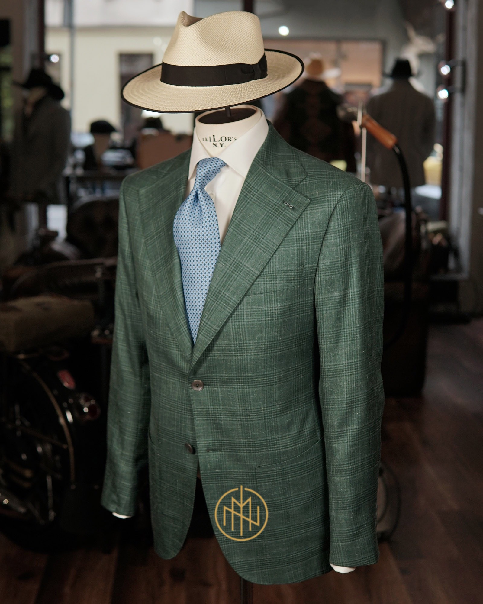Zielony, kraciasty garnitur na manekinie z kapeluszem i błękitnym krawatem, logo firmy na klapie, w tle wnętrze pracowni krawieckiej z antycznym pojazdem.