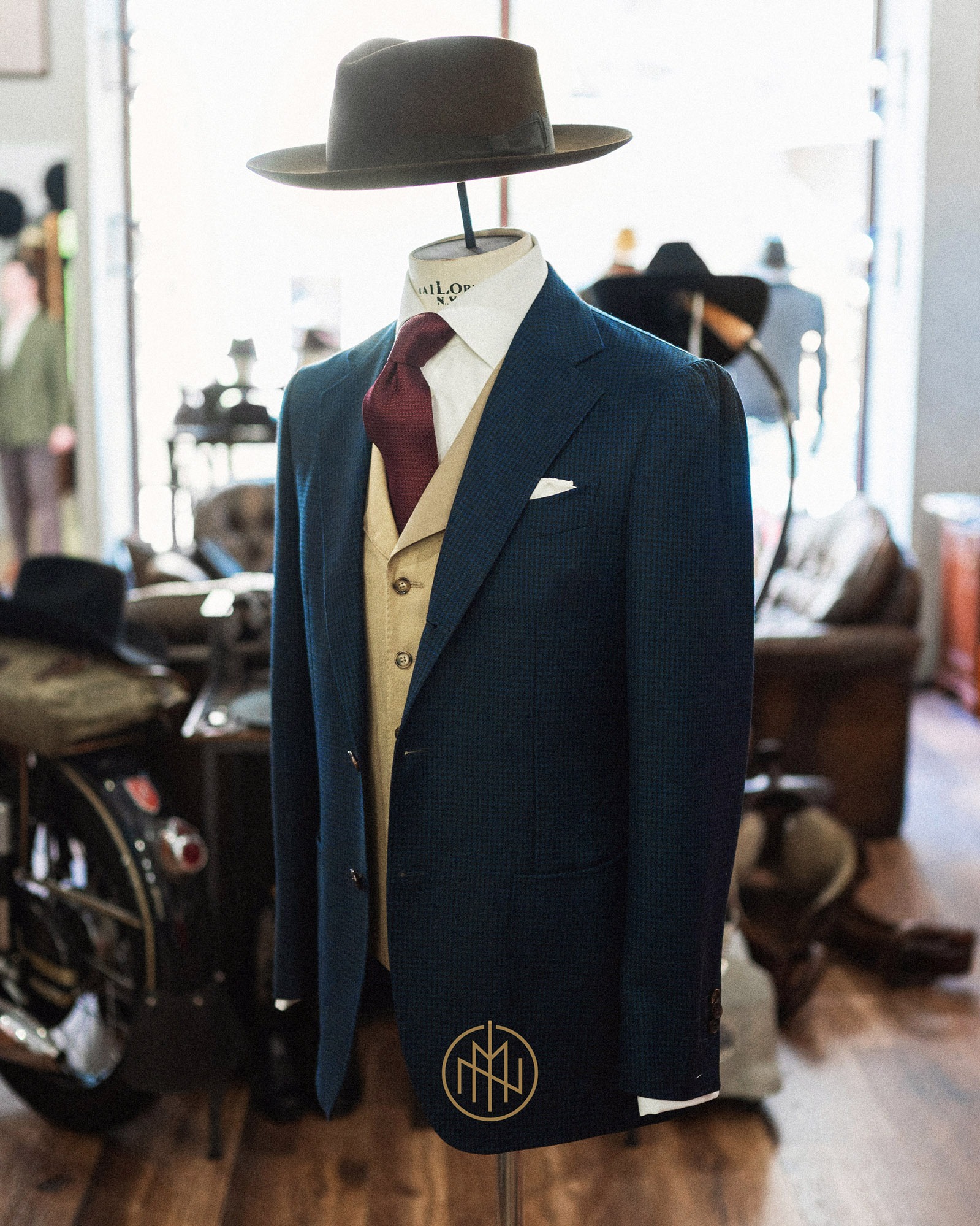Elegancki, granatowy garnitur z beżową kamizelką i bordowym krawatem na manekinie, uzupełniony brązowym kapeluszem. W tle widoczny warsztat krawiecki.