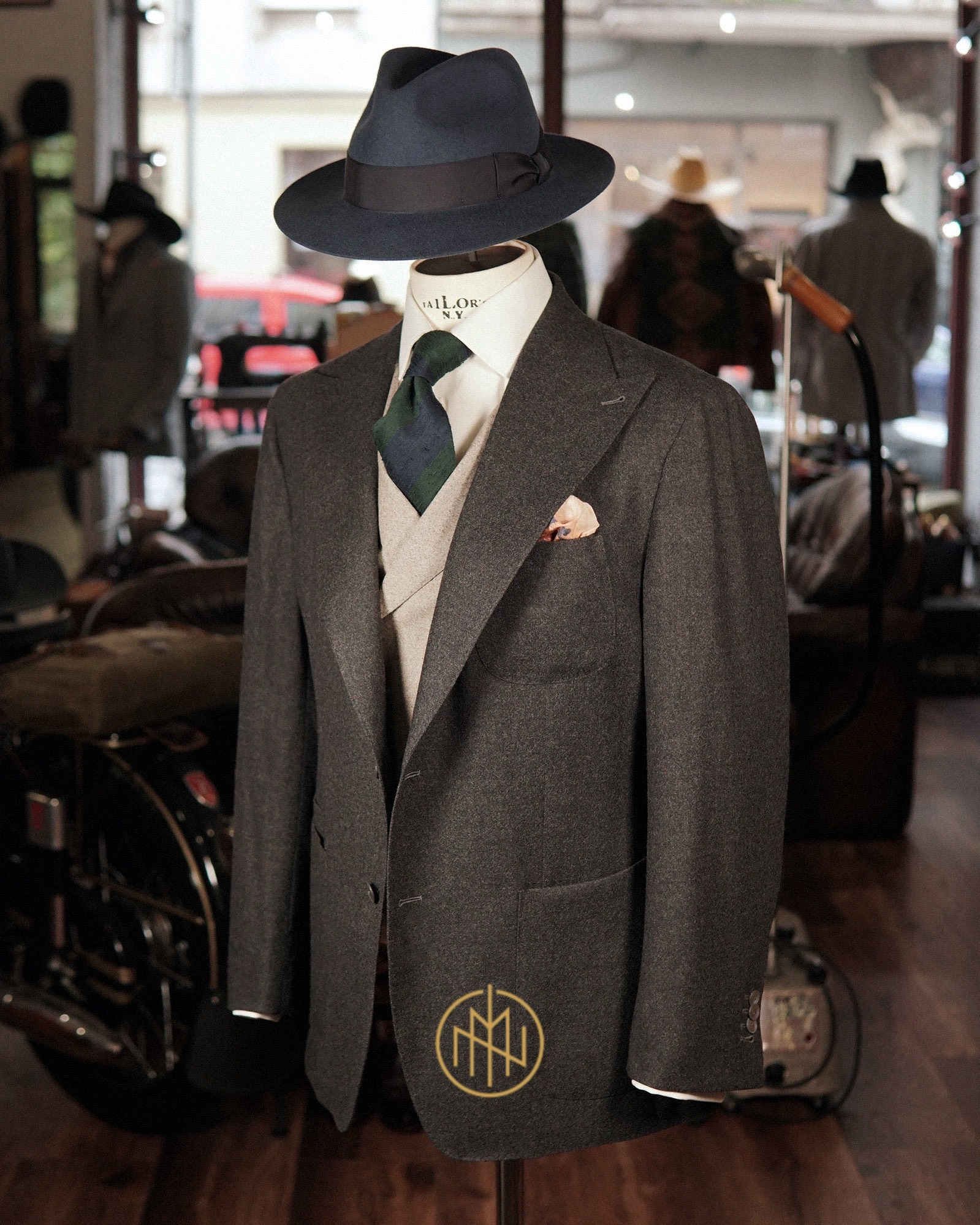 Elegancki, szary garnitur z wełny na manekinie, uzupełniony o kapelusz fedora, kamizelkę i krawat. W tle widoczny wystrój pracowni krawieckiej.
