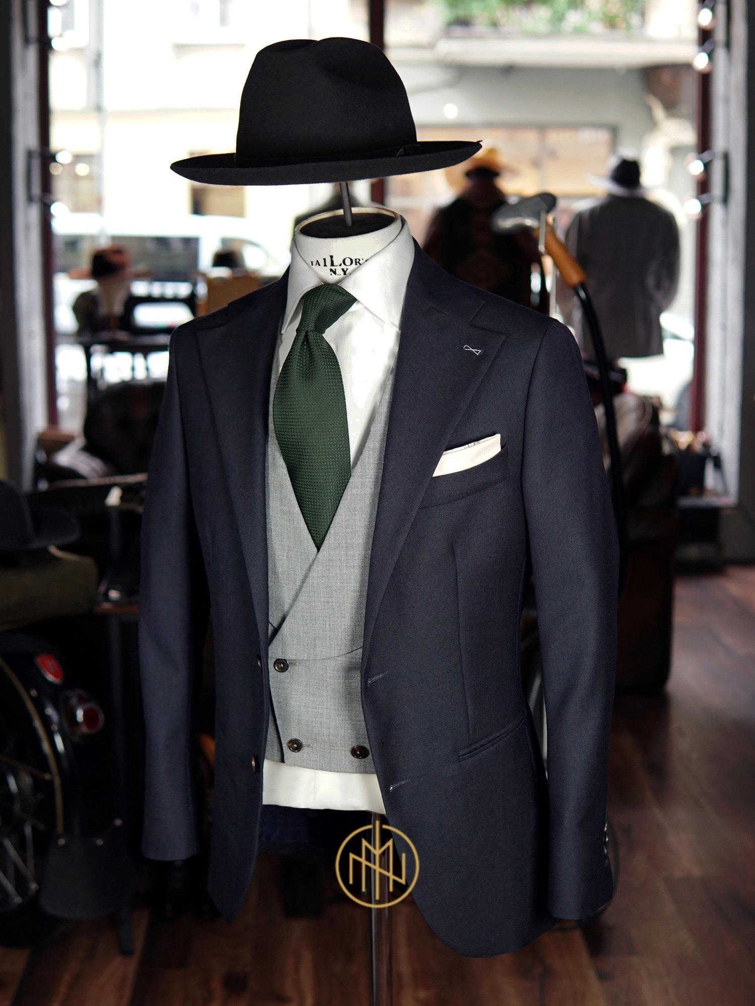 Elegancki, granatowy garnitur trzyczęściowy z zielonym krawatem i kapeluszem na manekinie w butiku krawieckim. Widoczne detale: poszetka, dwurzędowa kamizelka.