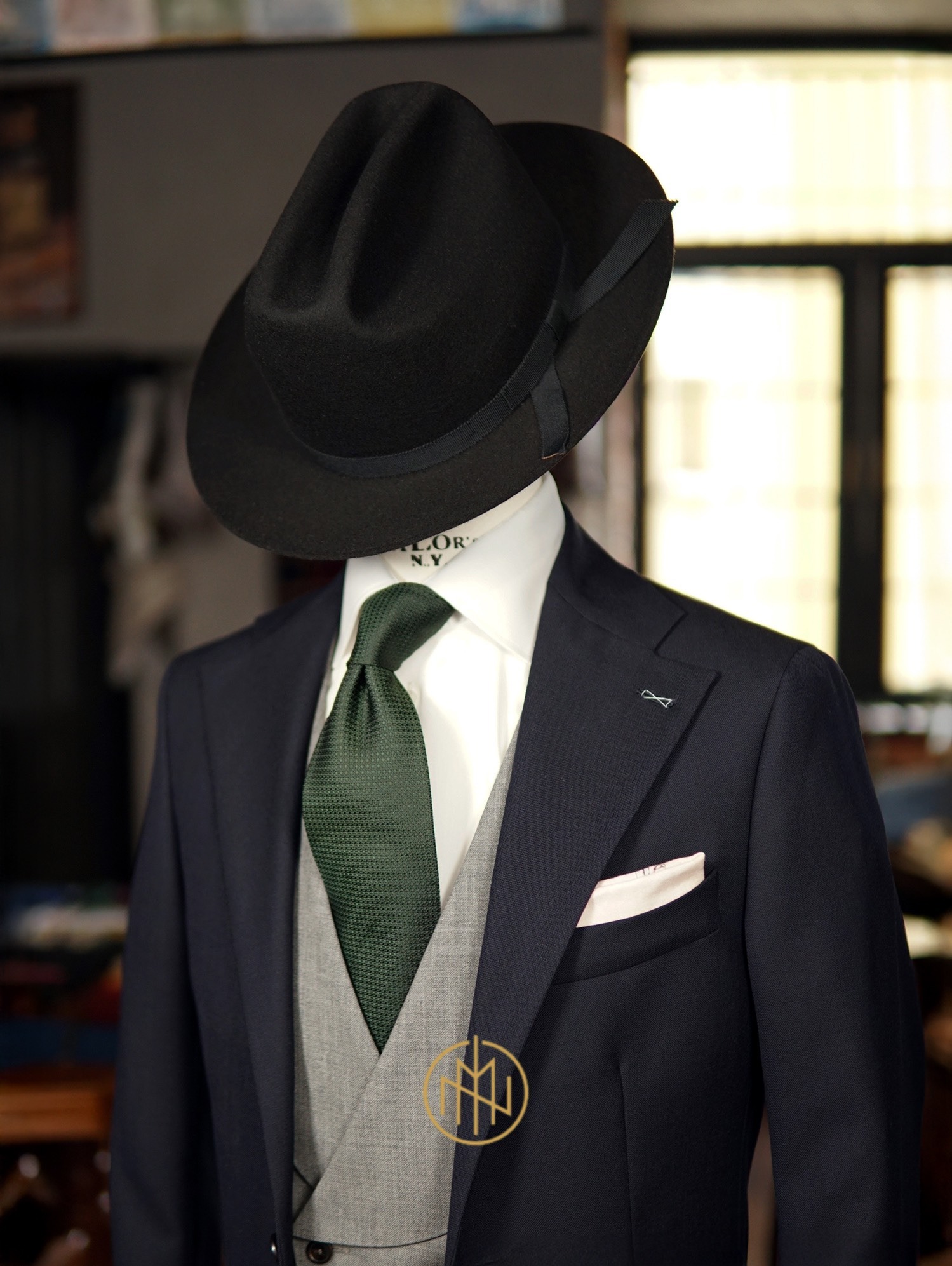 Elegancki, granatowy garnitur trzyczęściowy z zielonym krawatem i czarnym kapeluszem na manekinie. W kieszonce poszetka. Widoczne detale szycia.