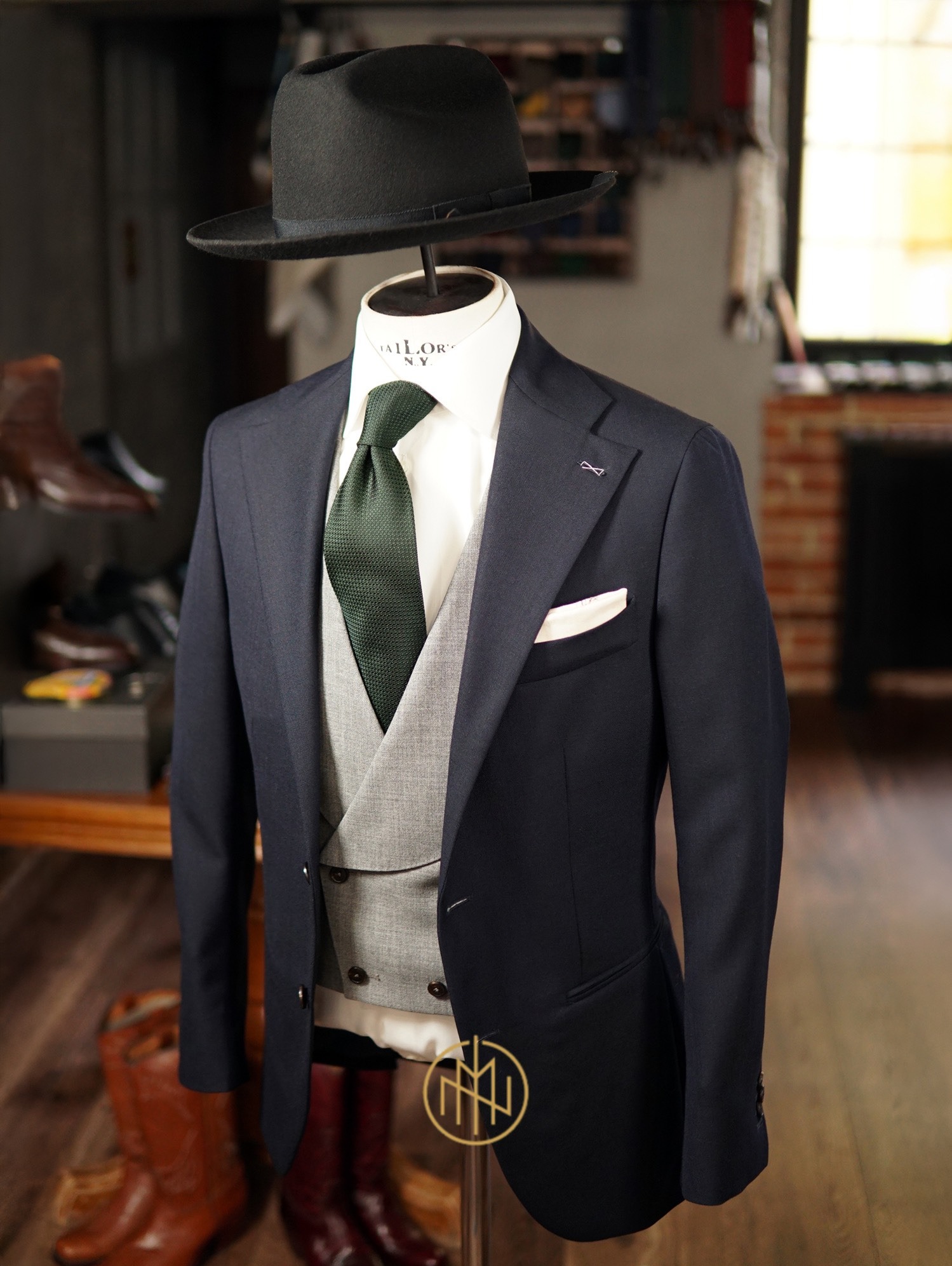 Elegancki, granatowy garnitur trzyczęściowy z szarą kamizelką, zielonym krawatem i kapeluszem na manekinie w pracowni krawieckiej. Widoczne buty i kominek w tle.