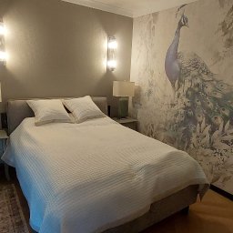 Kamil Dziupiński - Sypialnia z łóżkiem, szafkami nocnymi i lampami. Na ścianie tapeta z motywem pawia. Dwa kinkiety oświetlają ścianę. Widok zza futryny drzwi.