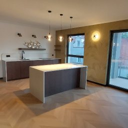 Kamil Dziupiński - Nowoczesne wnętrze z otwartą kuchnią, wyspą i drewnianą podłogą w jodełkę. Widoczne ciemne szafki, białe blaty, designerskie oświetlenie i duże okna balkonowe.