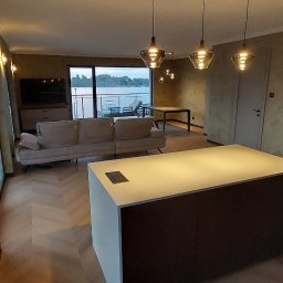 Kamil Dziupiński - Nowoczesny salon z aneksem kuchennym, jasną wyspą, podłogą w jodełkę i teksturowanymi ścianami w odcieniach beżu, z designerskimi lampami i widokiem na jezioro.