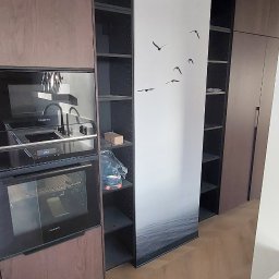 Kamil Dziupiński - Nowoczesna zabudowa kuchenna z ciemnego drewna, zintegrowanymi sprzętami Siemens i otwartymi półkami. Drewniana podłoga w jodełkę, dekoracyjna ściana z motywem morza i ptaków.