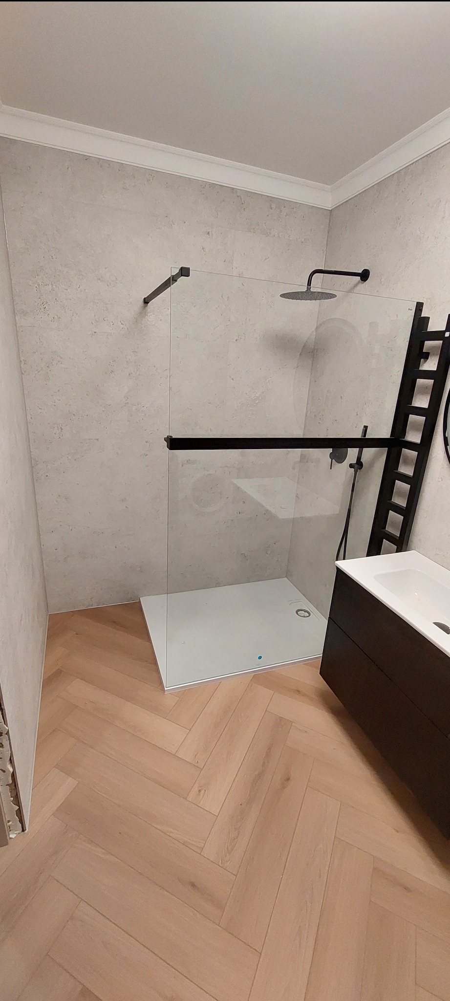 Nowoczesna łazienka z kabiną prysznicową typu walk-in, armaturą w kolorze czarnym i drewnianą podłogą ułożoną w jodełkę. Minimalistyczny design z elementami dekoracyjnymi.