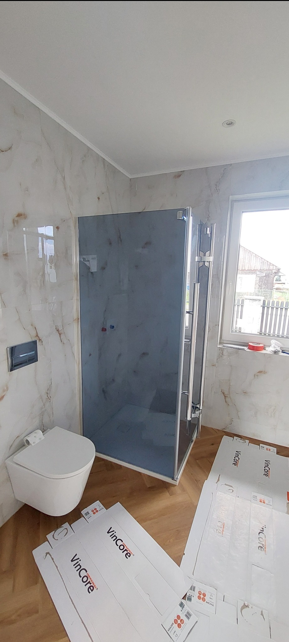 Wykończona łazienka z kabiną prysznicową, toaletą podwieszaną i marmurowymi płytkami. Na podłodze parkiet, a na podłodze leżą kartony po kabinie.
