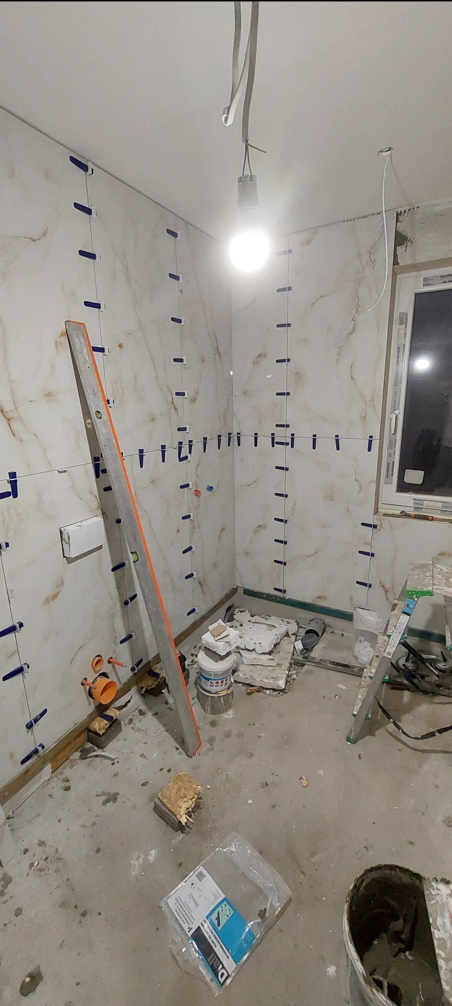 Układanie płytek imitujących marmur z użyciem poziomicy i klipsów w łazience w Kielcach. Widoczne narzędzia i materiały budowlane. Etap prac wykończeniowych.