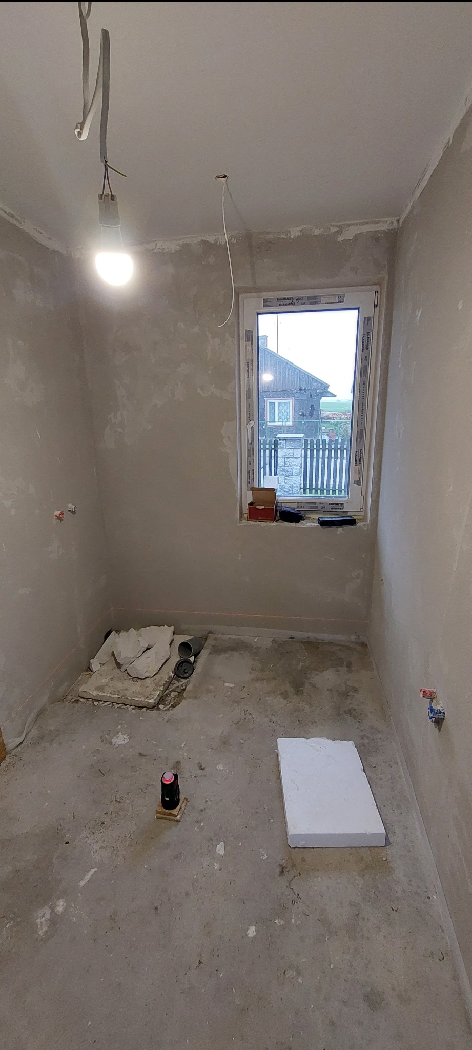 Surowy stan łazienki w trakcie prac: ściany po szpachlowaniu, widoczne instalacje, laser krzyżowy na podłodze, okno z widokiem na dom. Etap przygotowania do dalszych prac.