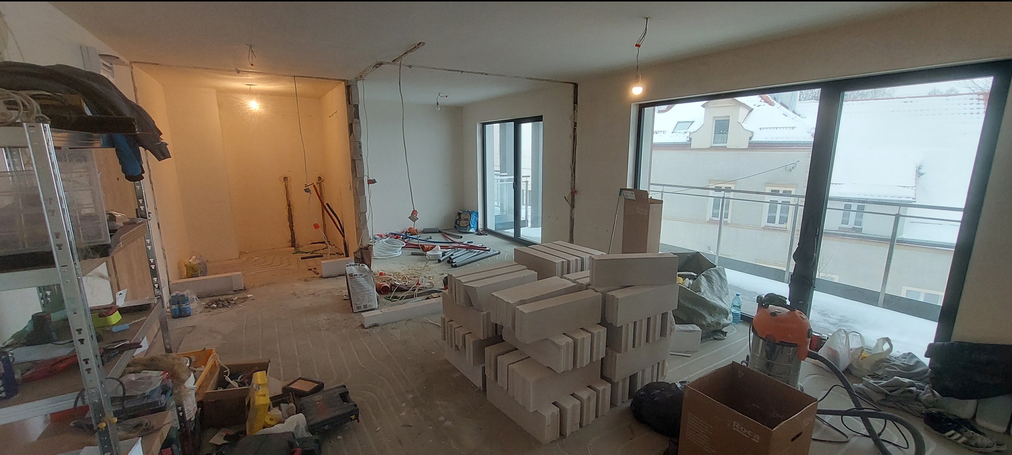 Wnętrze w trakcie remontu: widoczne materiały budowlane, instalacje i narzędzia. Prace przygotowawcze przed wykończeniem, z widokiem na zaśnieżony krajobraz za oknem.