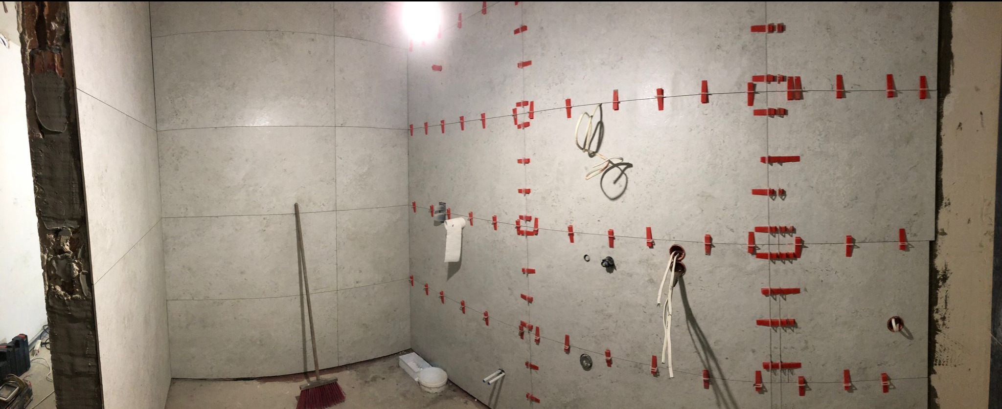 Ściany łazienki w trakcie układania płytek, z widocznymi systemami poziomowania (czerwone klipsy i sznurki). Fragmenty instalacji elektrycznej i hydraulicznej.