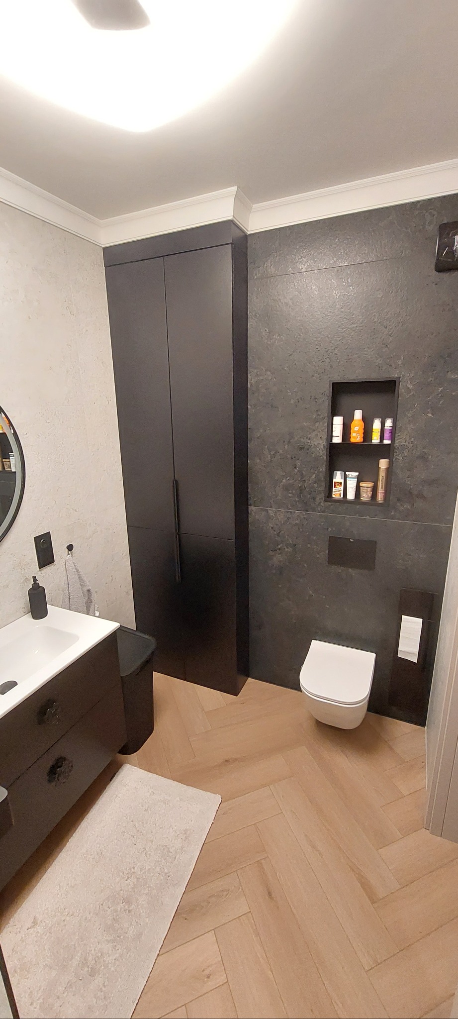 Nowoczesna łazienka z drewnopodobną podłogą w jodełkę, czarną szafką umywalkową z ozdobnymi uchwytami i wnęką w ścianie z kosmetykami.