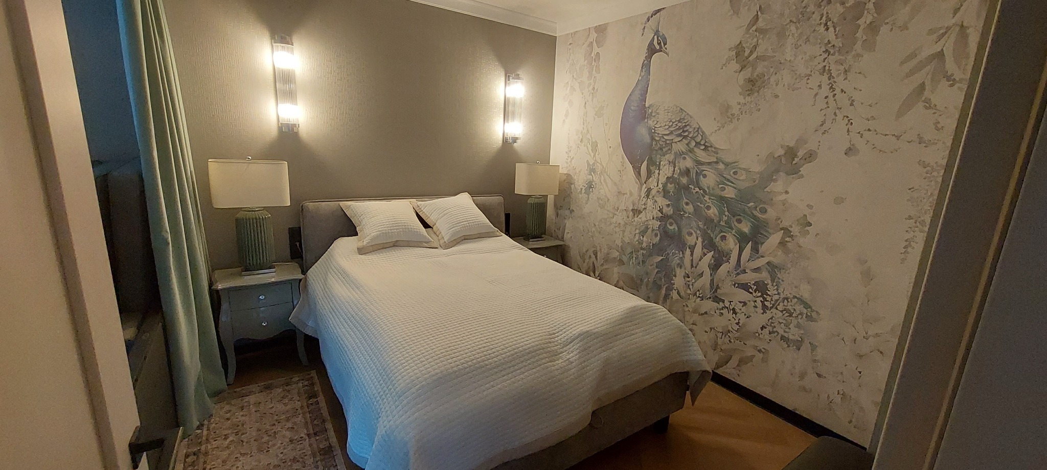 Sypialnia z łóżkiem, szafkami nocnymi i lampami. Na ścianie tapeta z motywem pawia. Dwa kinkiety oświetlają ścianę. Widok zza futryny drzwi.