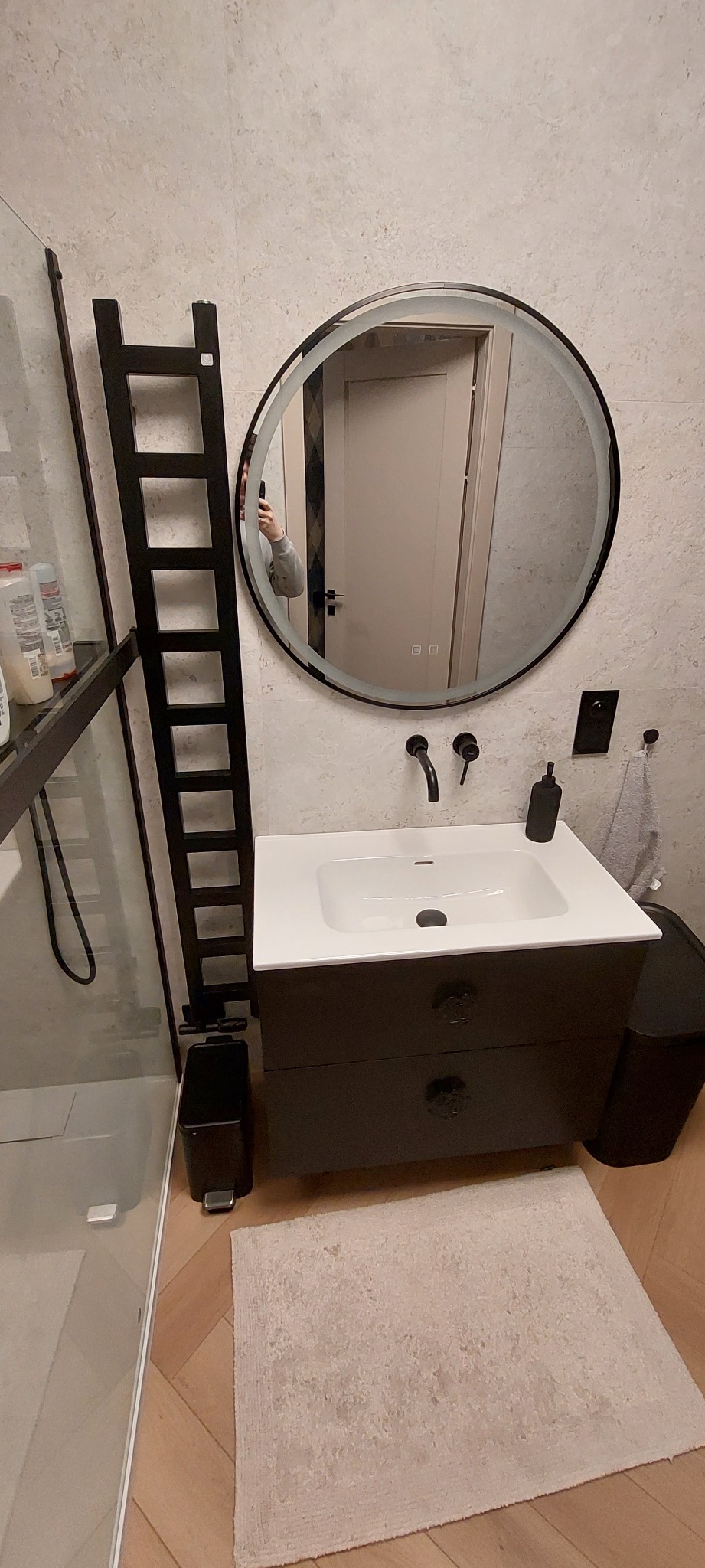 Nowoczesna łazienka z okrągłym lustrem z podświetleniem LED, czarną armaturą i drabinką na ręczniki. Jasne płytki i drewniana podłoga tworzą spójną całość.