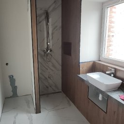 Azariasz Janicki ZIAJ-TECH - Nowoczesna łazienka w trakcie wykończenia: białe płytki imitujące marmur, drewniane panele ścienne, kabina prysznicowa i umywalka w oknie. Widoczne ślady prac wykończeniowych.