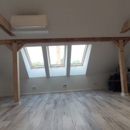 Azariasz Janicki ZIAJ-TECH - Jasne poddasze po remoncie z drewnianymi belkami, trzema oknami dachowymi i panelami podłogowymi imitującymi drewno. Widoczna drabina i odkurzacz.